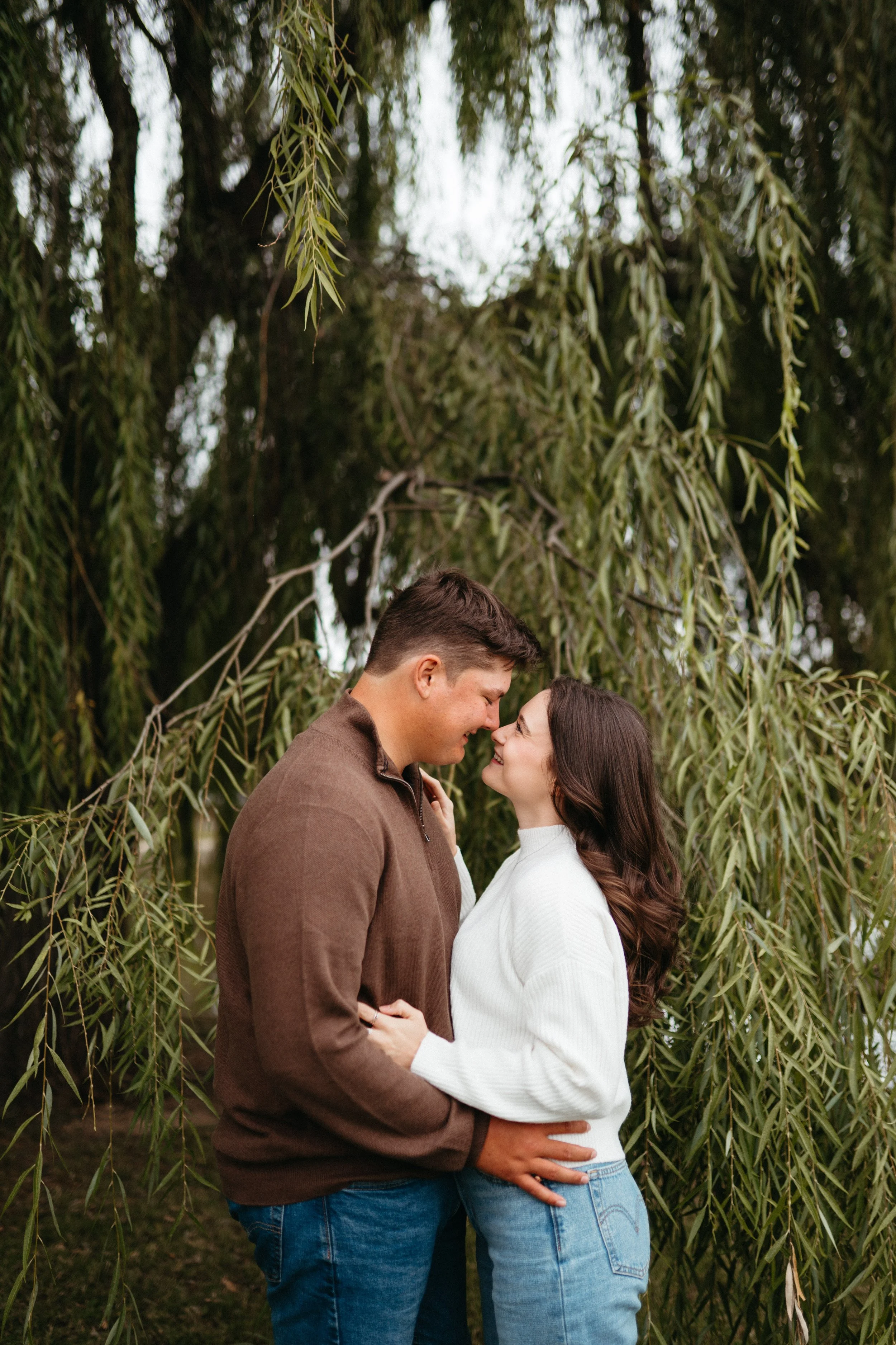 A couple standing close together, touching noses, and smiling in front of hanging tree branches.