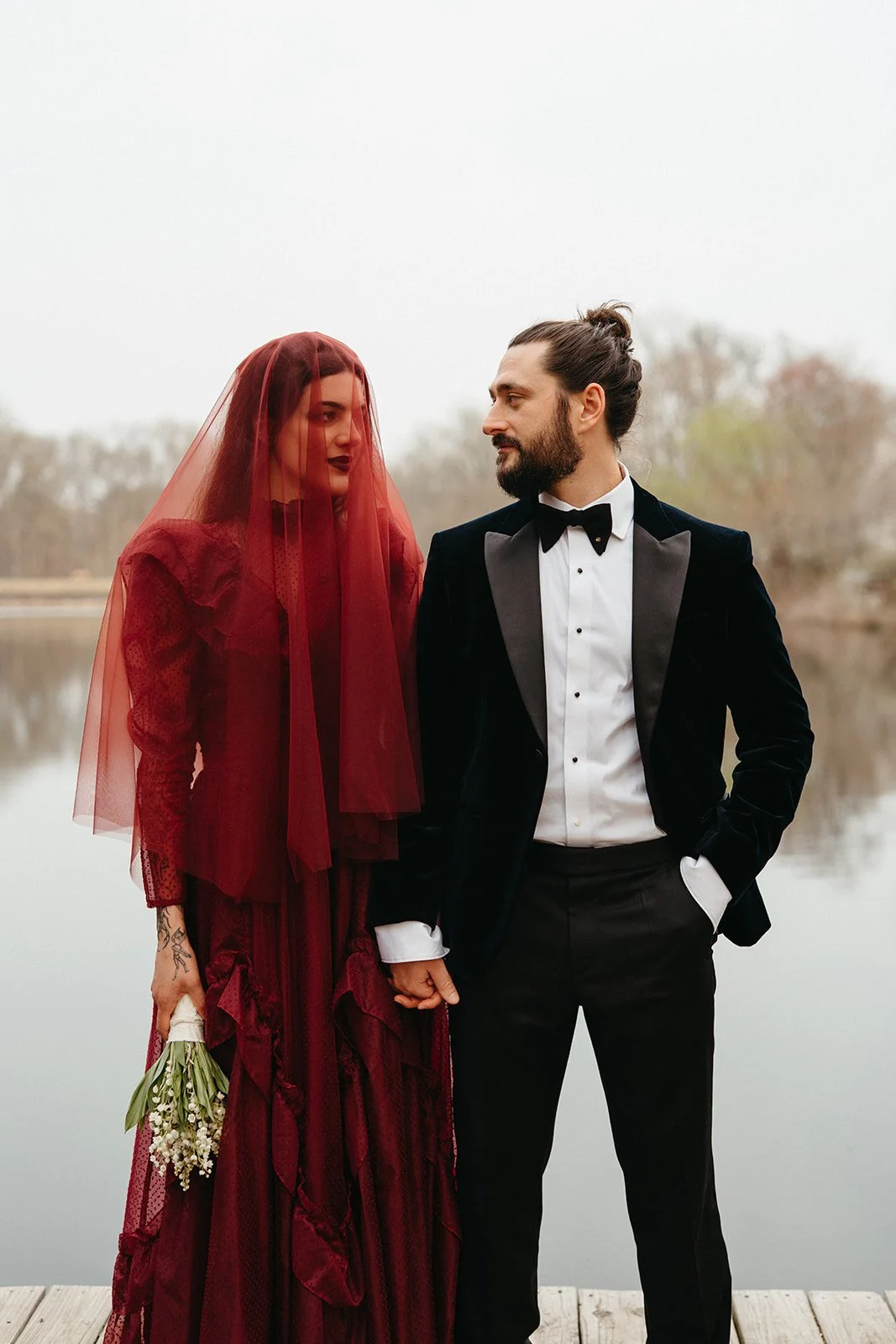 A couple dressed in wedding attire standing near a lake, holding hands, with the woman wearing a red dress and veil, and the man in a black tuxedo, during daytime.
