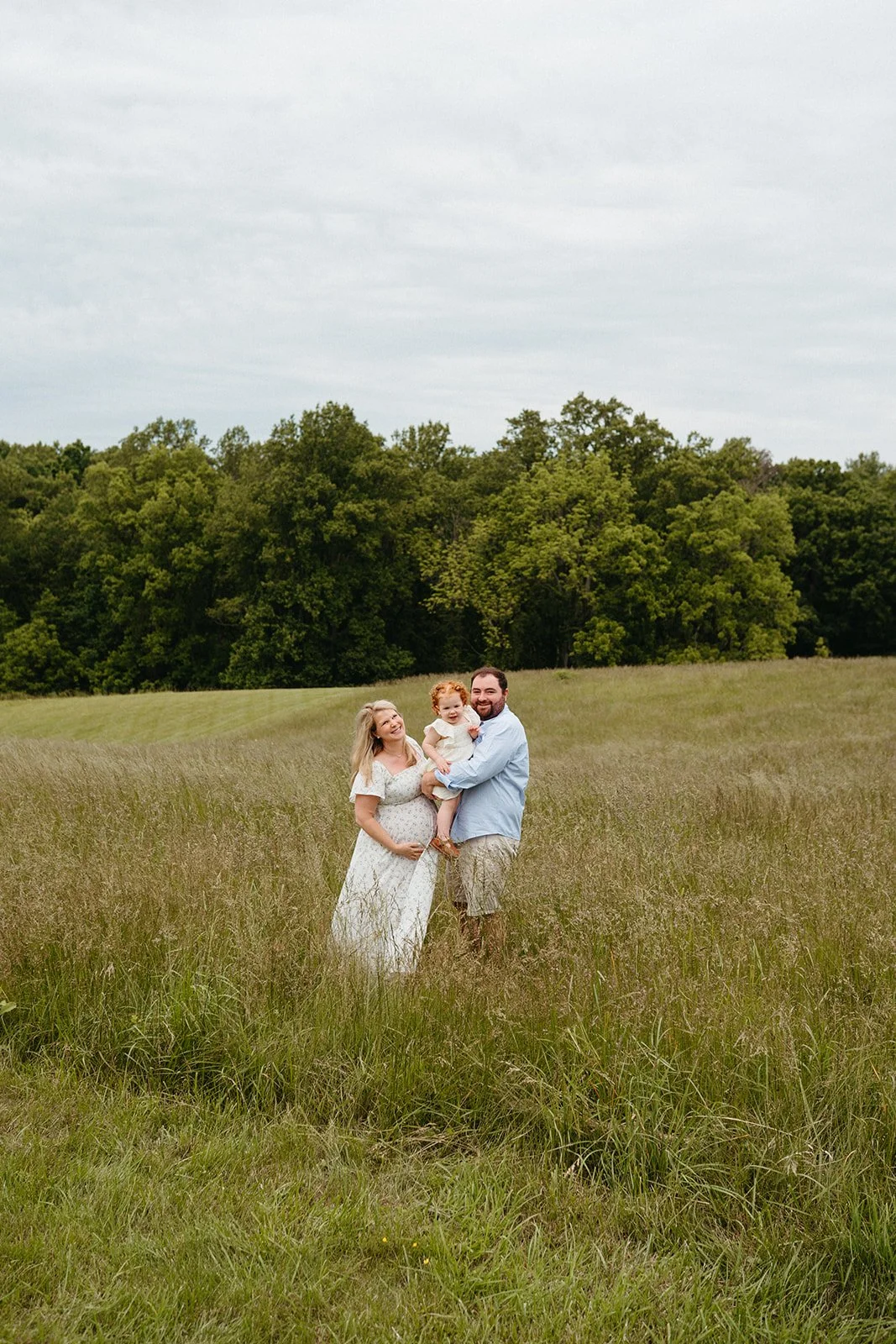 A family of three standing in a grassy field with trees in the background, smiling and enjoying a sunny day.