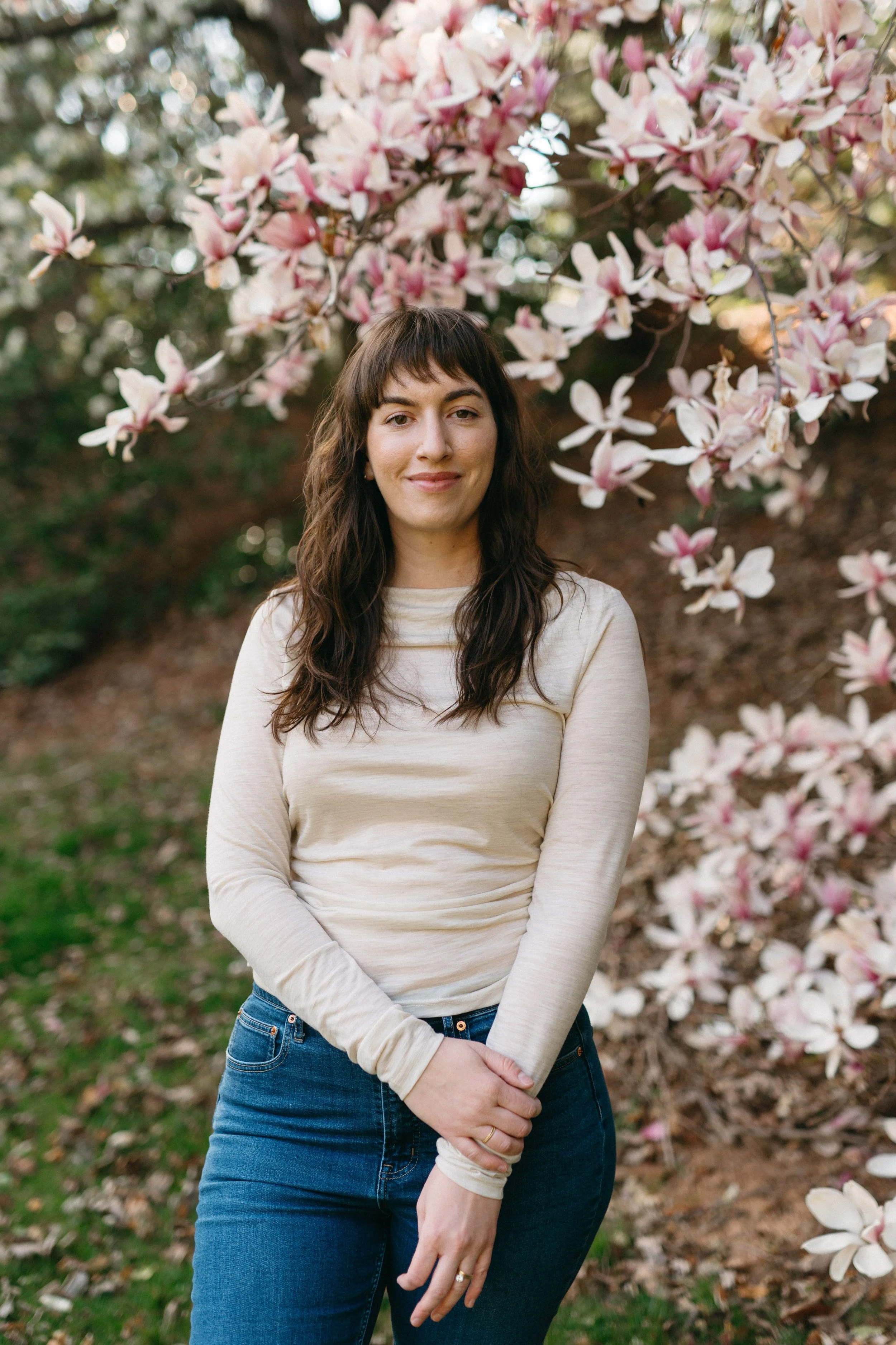 A woman with long wavy brown hair, wearing a light-colored long sleeve shirt and blue jeans, standing outdoors in front of a magnolia tree with pink and white blossoms.