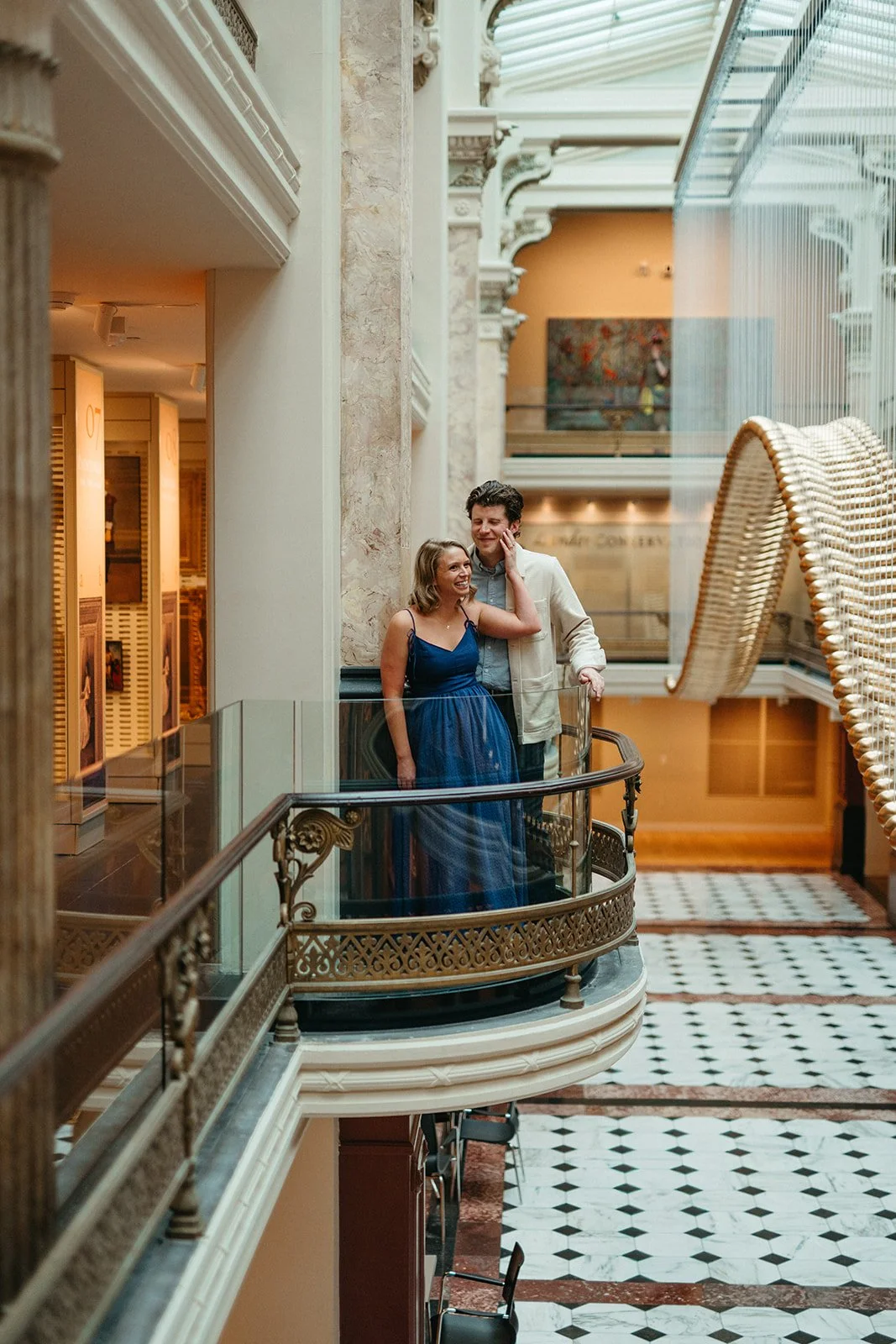 A young couple standing on a decorative balcony inside a museum or historic building, smiling and enjoying each other's company.
