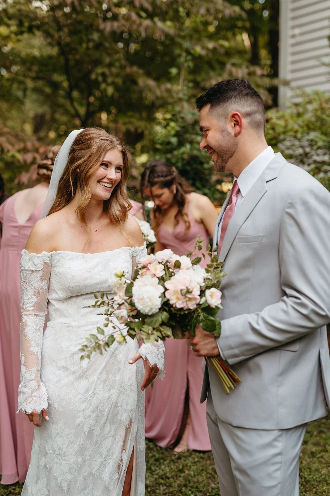 A bride and groom smiling at each other during their outdoor wedding, with bridesmaids in pink dresses in the background.