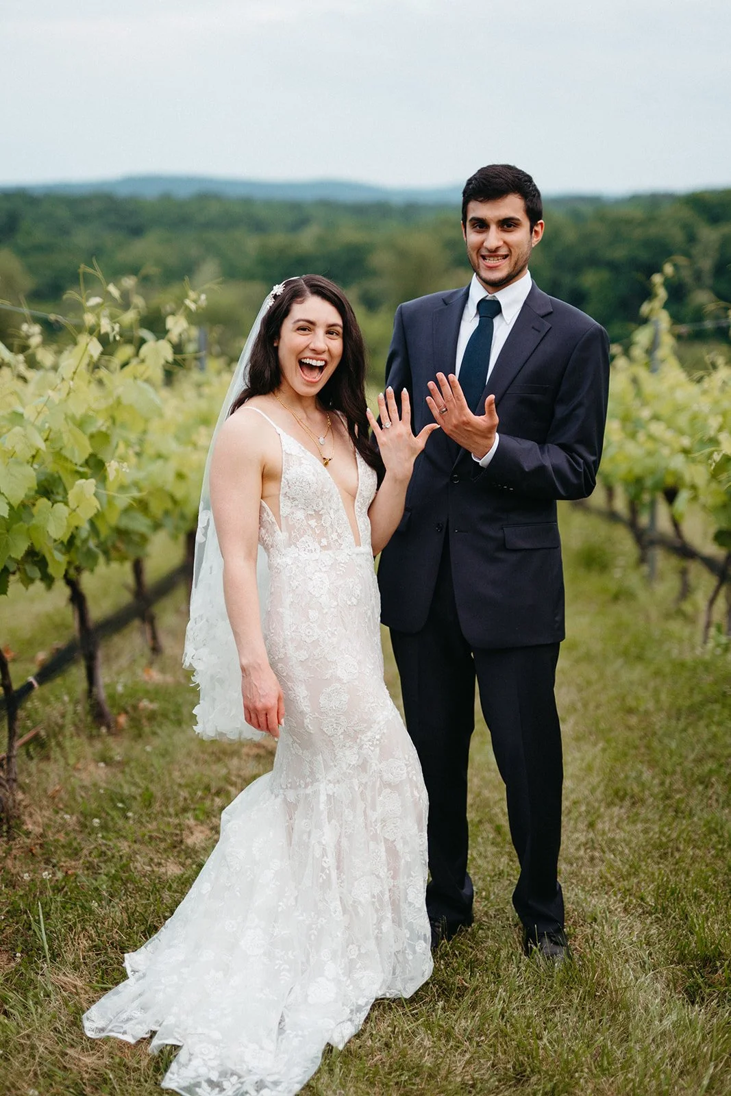 A joyful bride and groom showing off their wedding rings in a vineyard with green hills in the background.