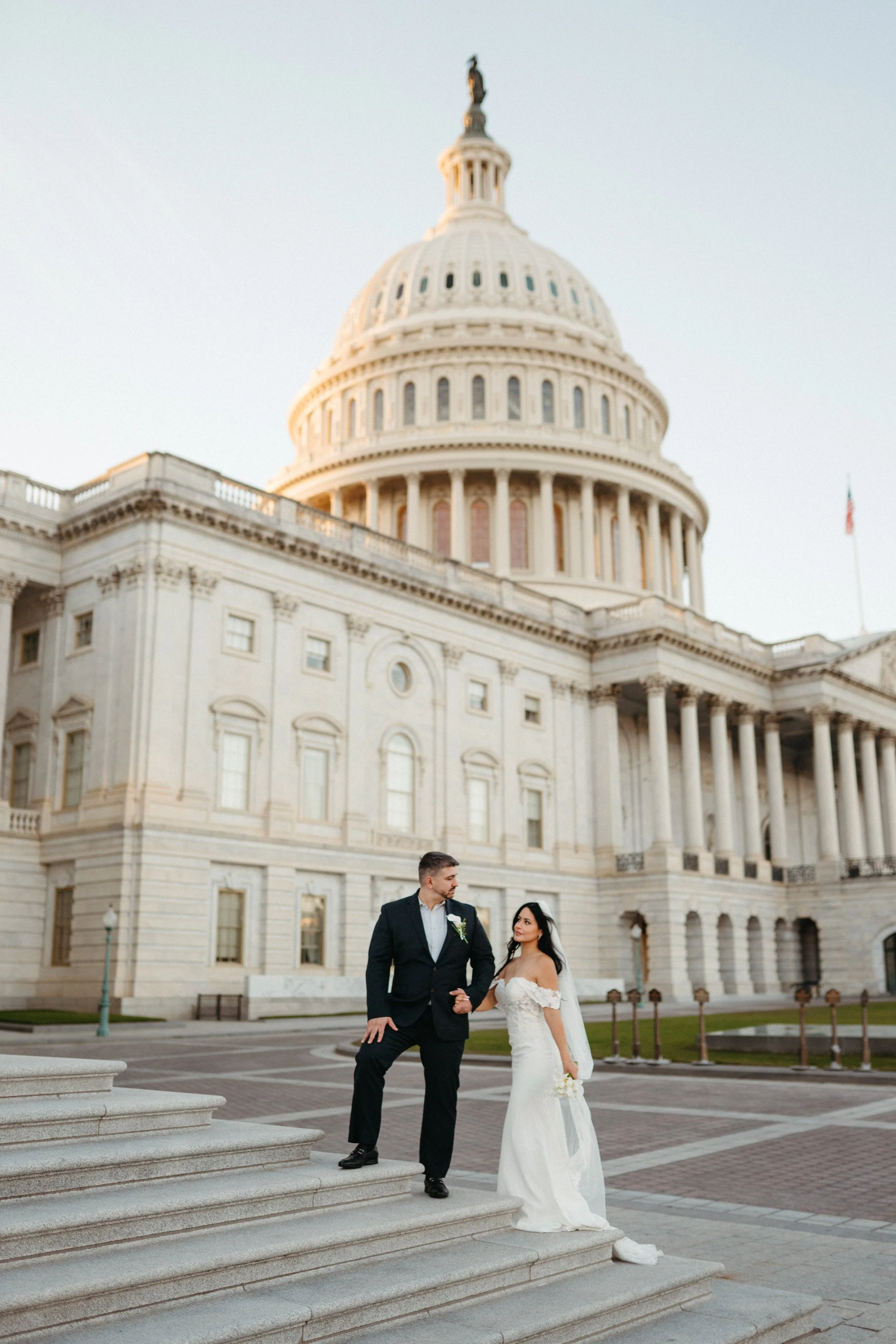 A newlywed couple dressed in wedding attire standing on stairs in front of the United States Capitol building in Washington, D.C.