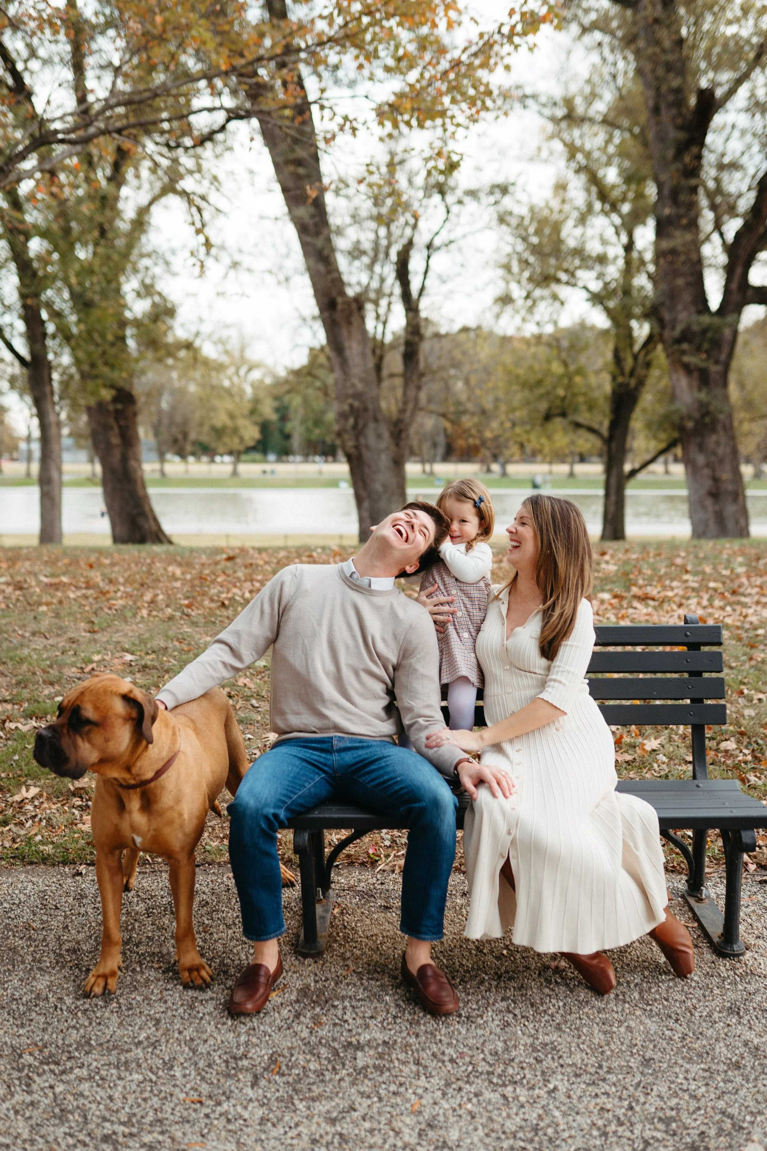 A happy family of three and a dog sitting on a park bench in autumn. The parents and daughter are laughing and enjoying the outdoor setting with fall leaves and trees in the background.