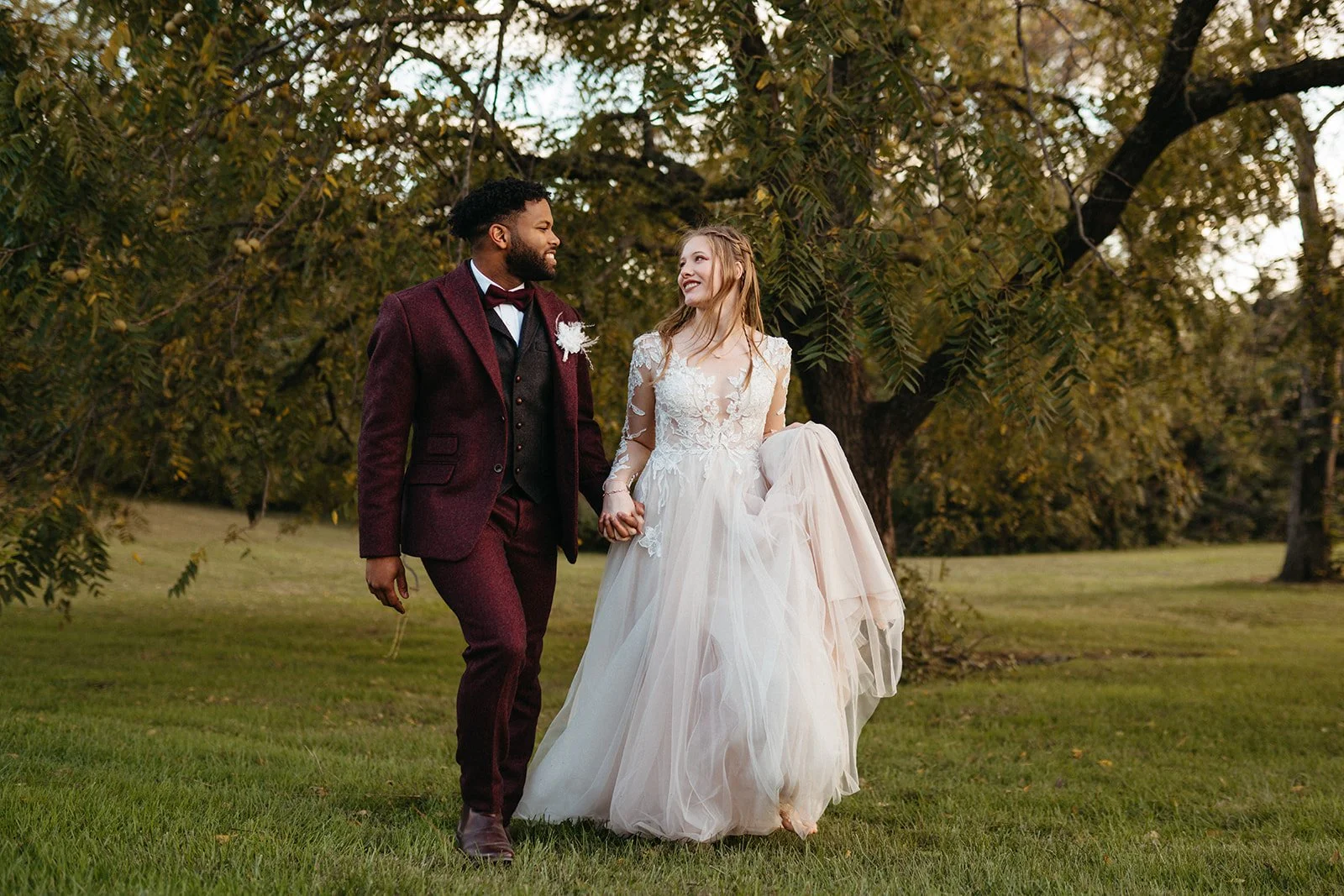 A newlywed couple walking hand in hand on a grassy field with trees in the background, smiling at each other during a photo shoot.