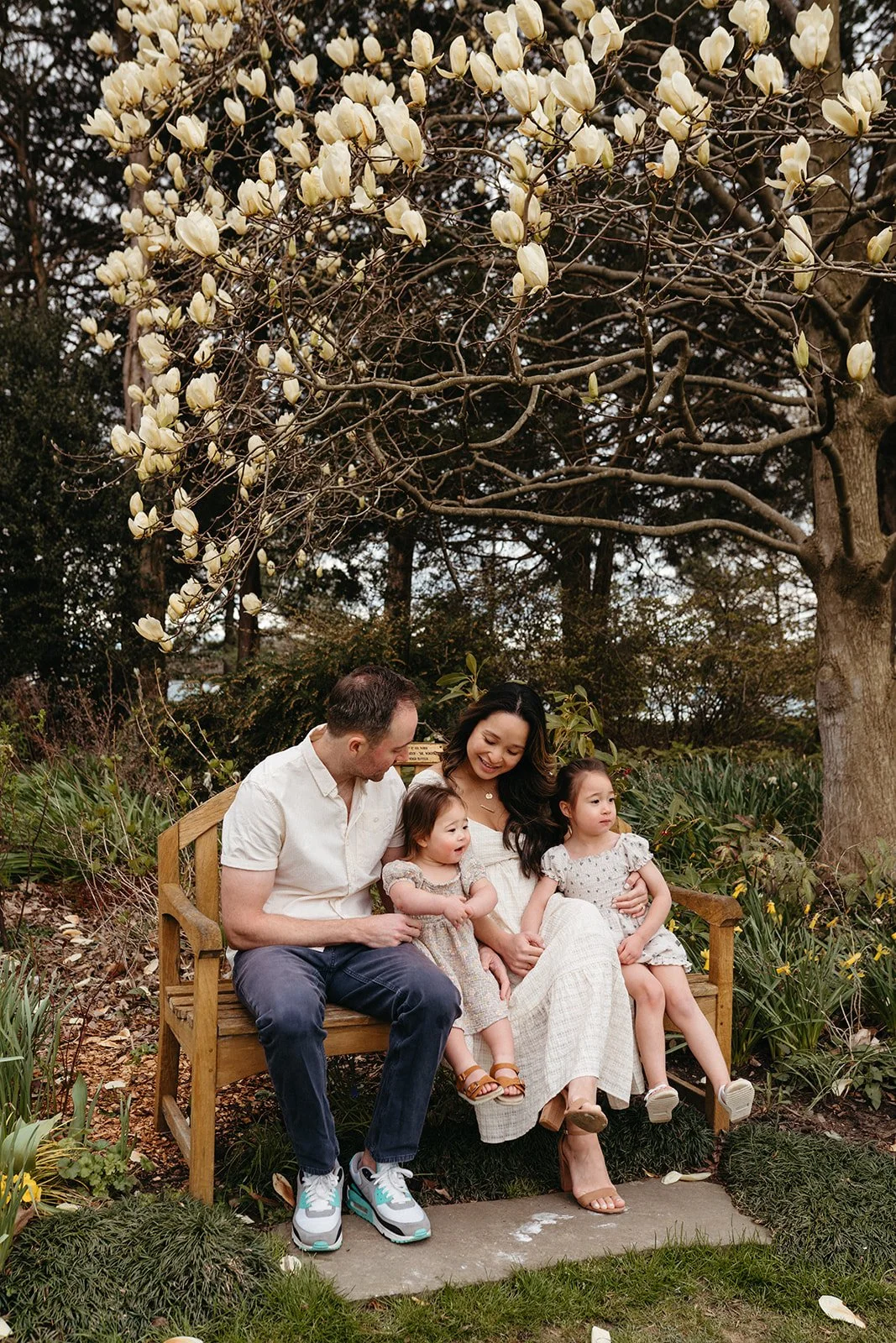 A family of four sitting on a wooden bench outdoors beneath a flowering tree, enjoying a moment together. The mother and father are smiling at their two young daughters, who are sitting on their laps.