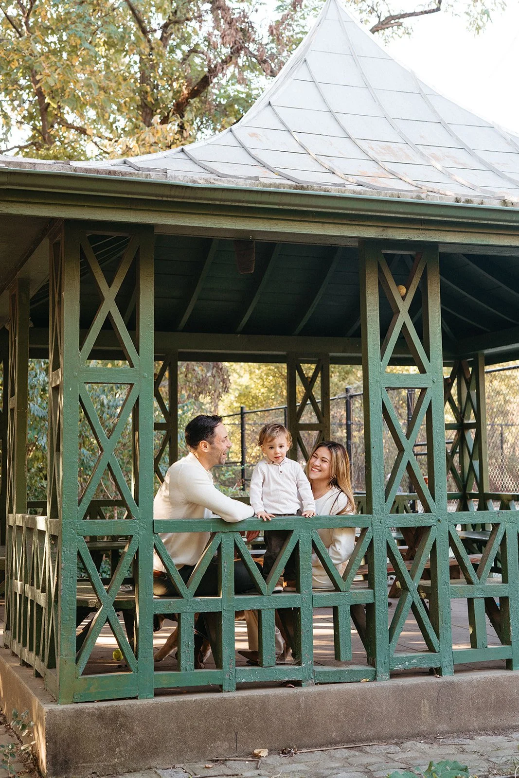 A family of three—father, mother, and young son—smiling and enjoying time together in a green wooden gazebo outdoors. The father and mother are sitting at the sides, and the son stands between them, all smiling and facing each other.