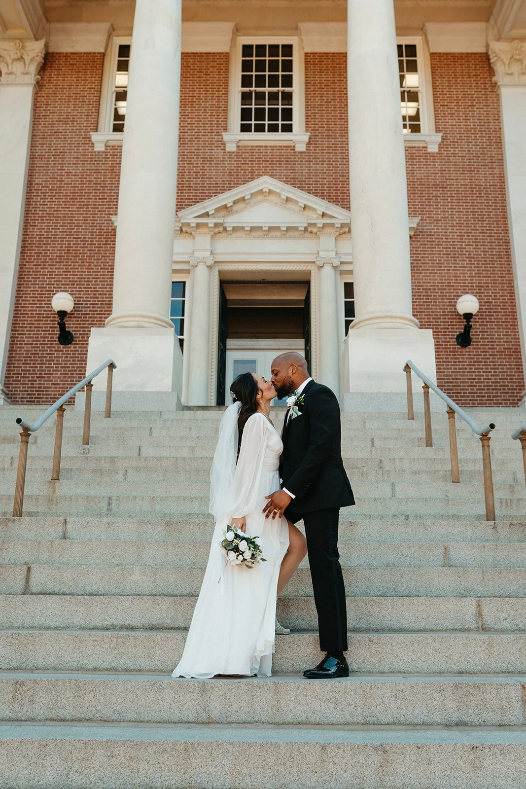 A bride and groom sharing a kiss on the steps of a large building with tall columns and brick walls.