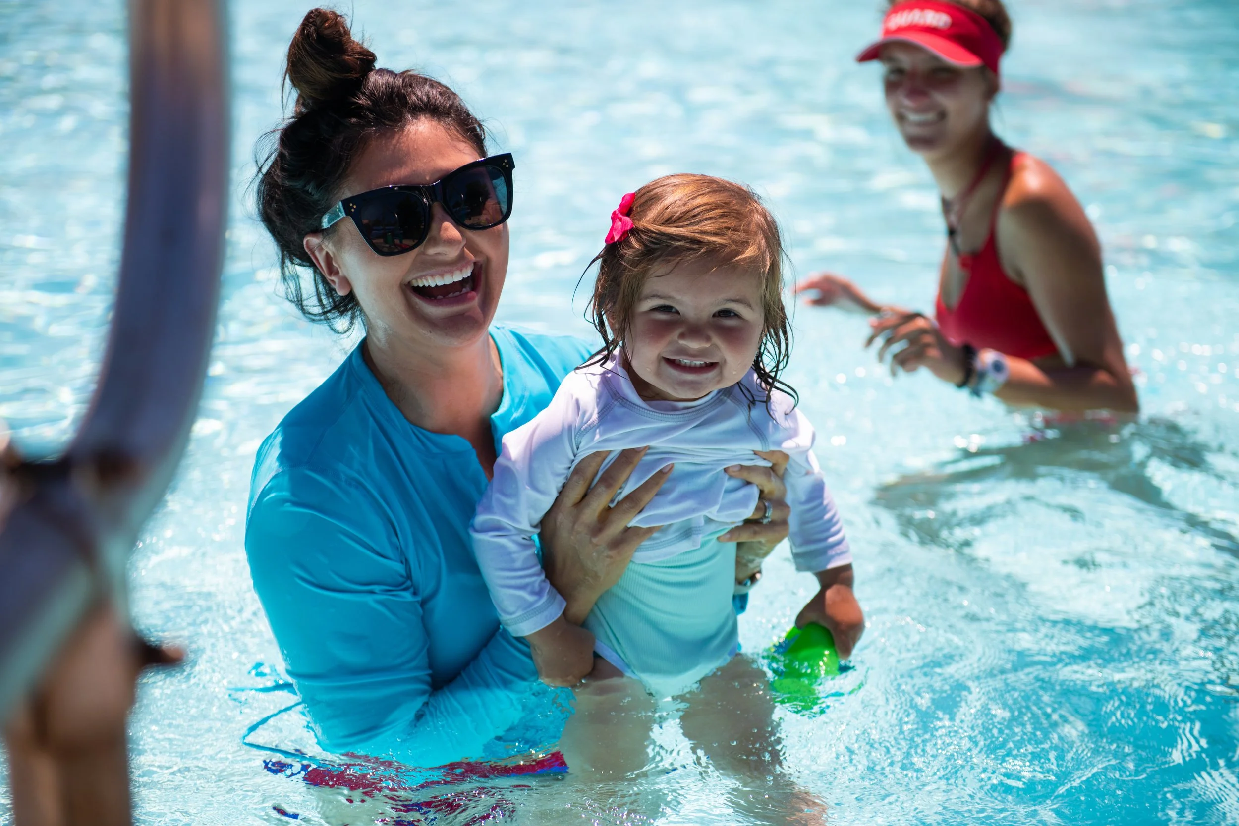 Three women and a young girl enjoying a swim in a pool. The woman in the foreground is wearing sunglasses and a blue shirt, holding the girl who is smiling and holding green floaties, with two other women smiling and playing in the background.