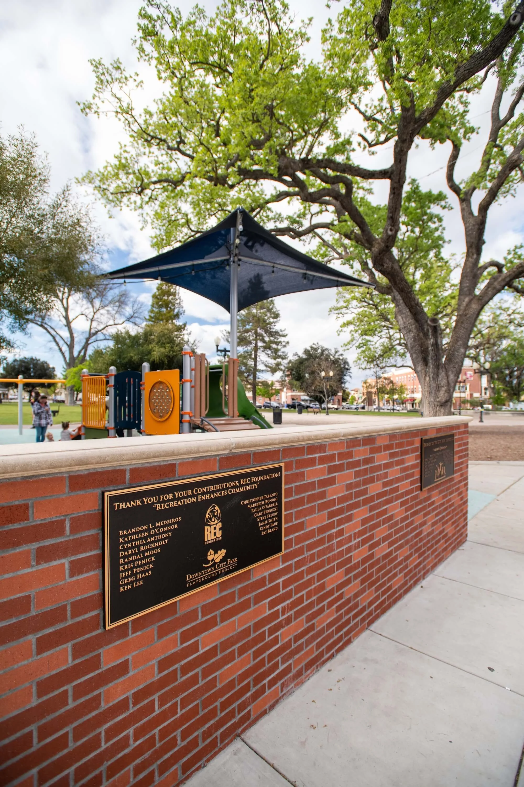A playground with a large tree, a blue shade canopy, and colorful play structures in a park. There is a brick wall with plaques displaying donations and acknowledgments.