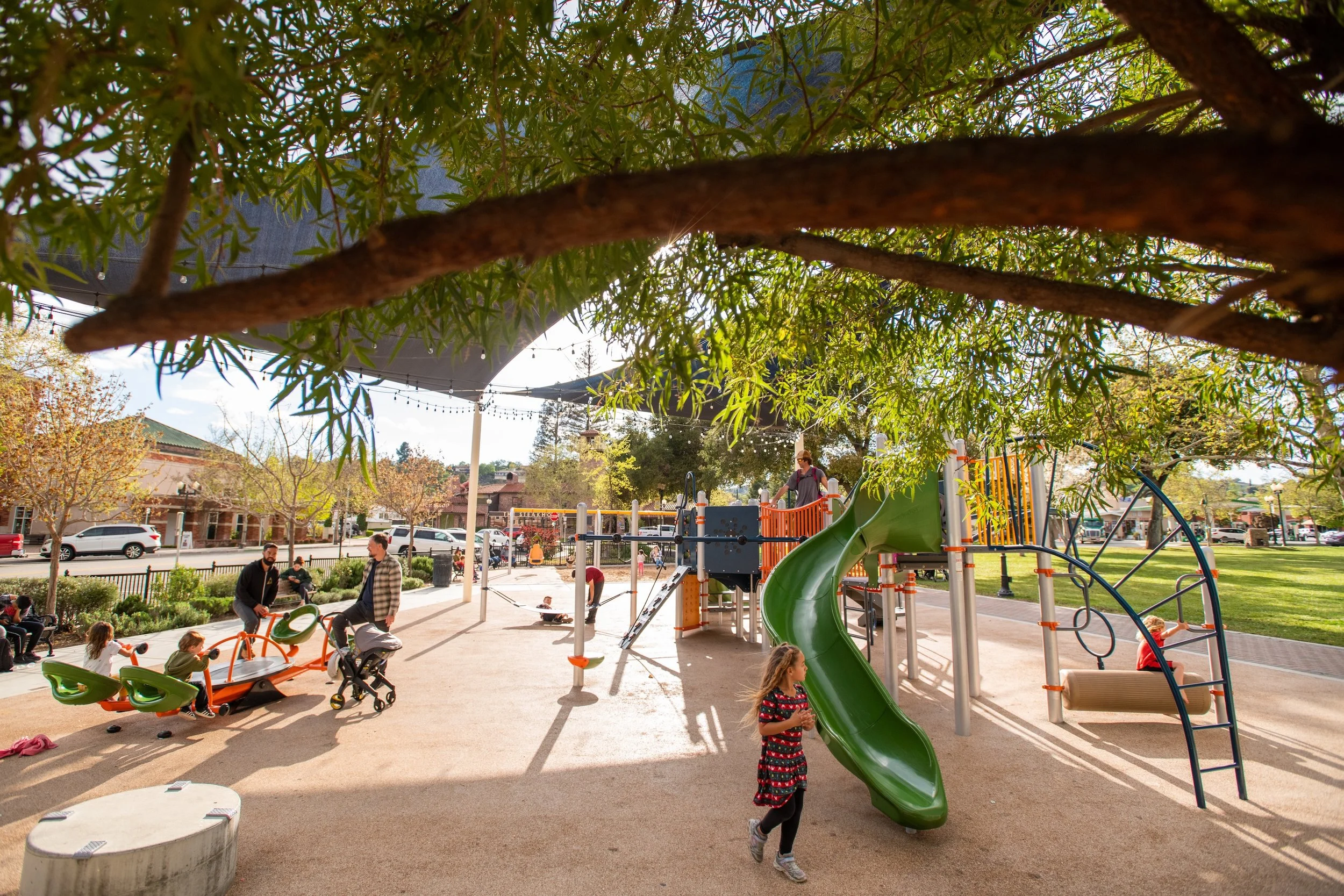 Children playing at a park with a green slide, a seesaw, and swing, under a large shade canopy, with adults and other children in the background, trees lining the park