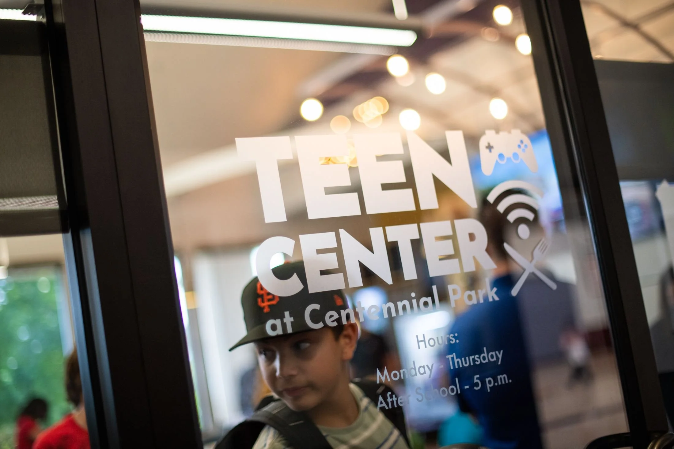 Kids playing at Teen Center in Centennial Park, looking through glass door with hours listed.