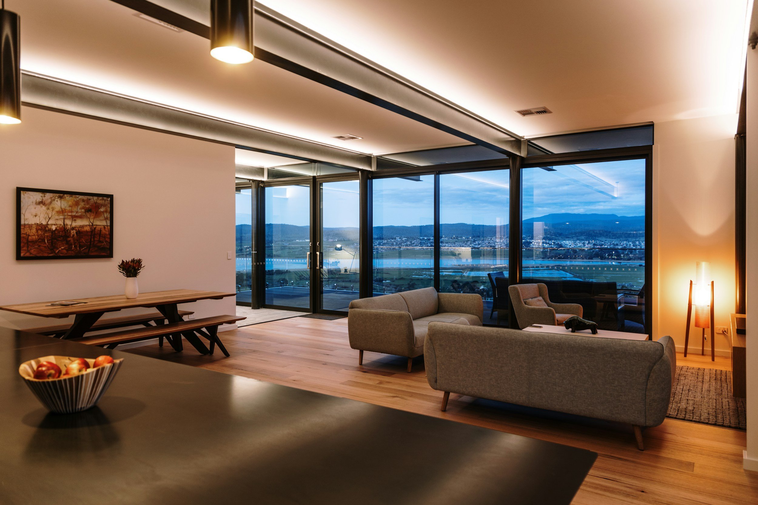 Modern living room with large windows showing a city view, light wood flooring, beige upholstered sofas, a wooden dining table with benches, a bowl of apples, and a floor lamp with a warm glow. Demonstrating lighting and outlet installation.