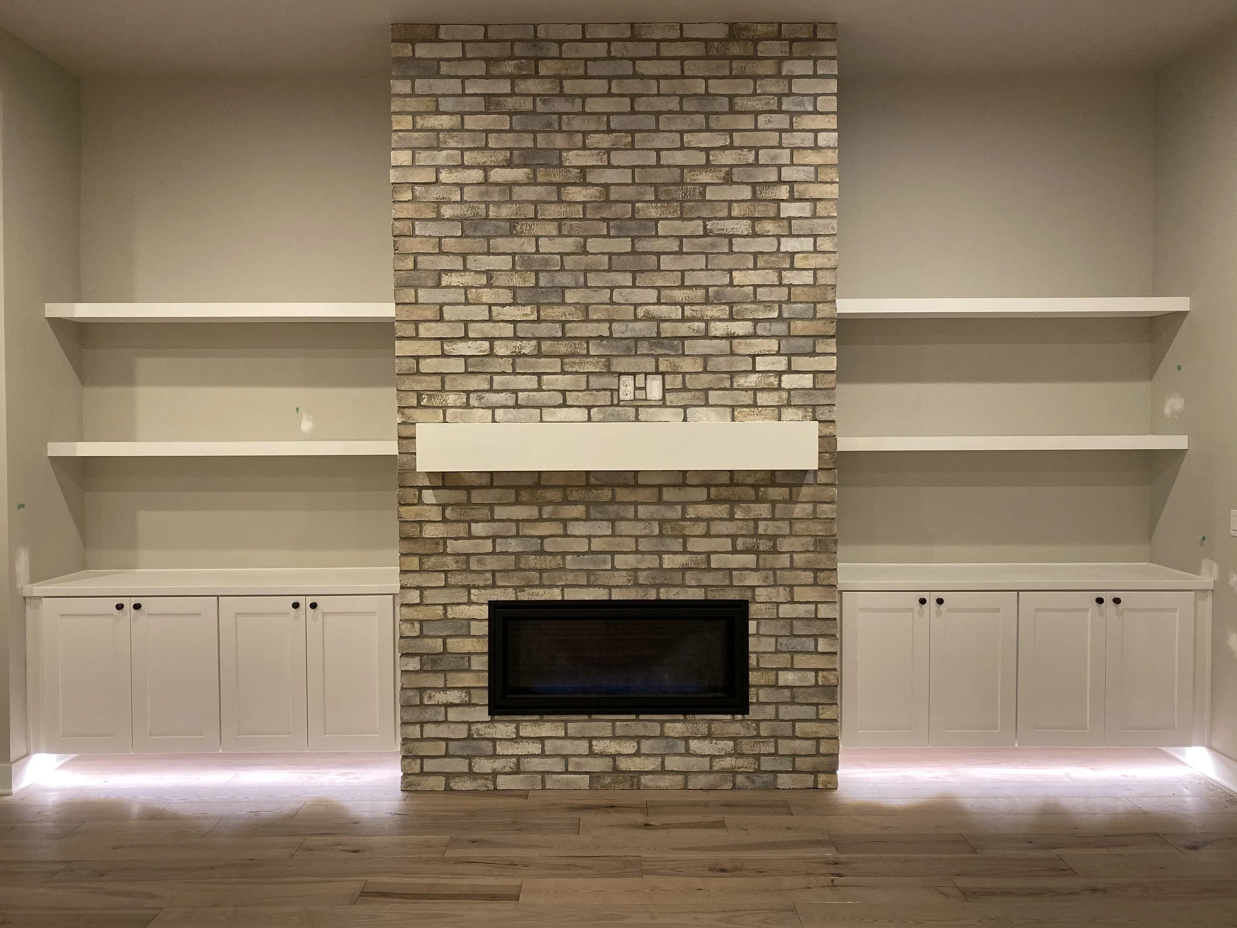 Empty white built-in shelves with cabinets beneath, flanking a brick fireplace with a mantel, in a living room with wood flooring.