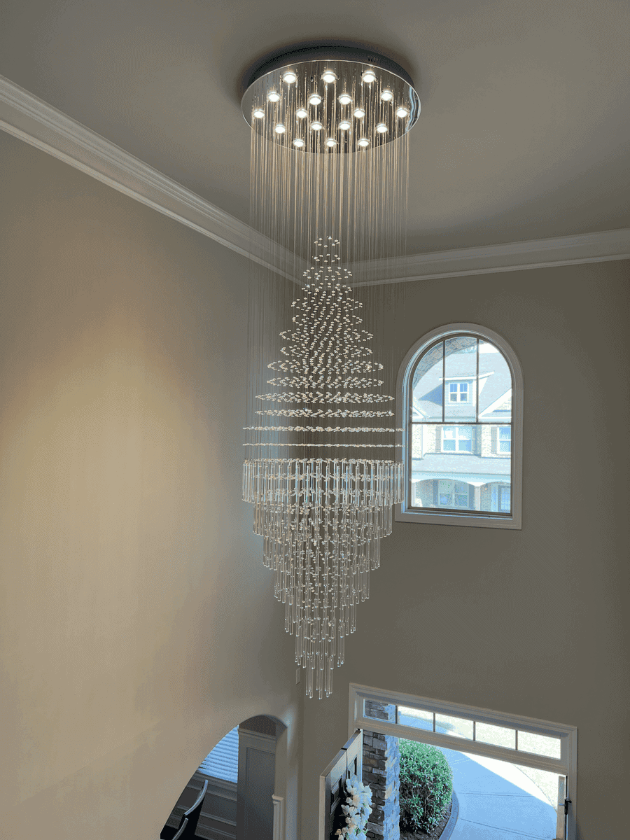 Large crystal chandelier hanging from a ceiling in a home interior with a high vaulted ceiling, a window, and a partial view of the front door area. Demonstrating lighting and electrical fixture installation and repair services.