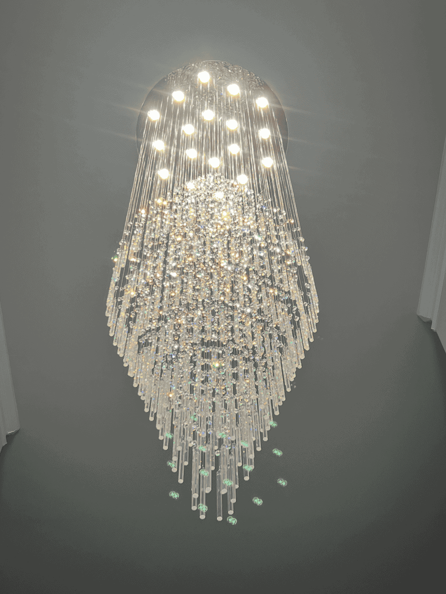 A large, elegant chandelier with multiple tiers of hanging crystal prisms and bright lights at the top. Demonstrating luxury indoor lighting installation. 
