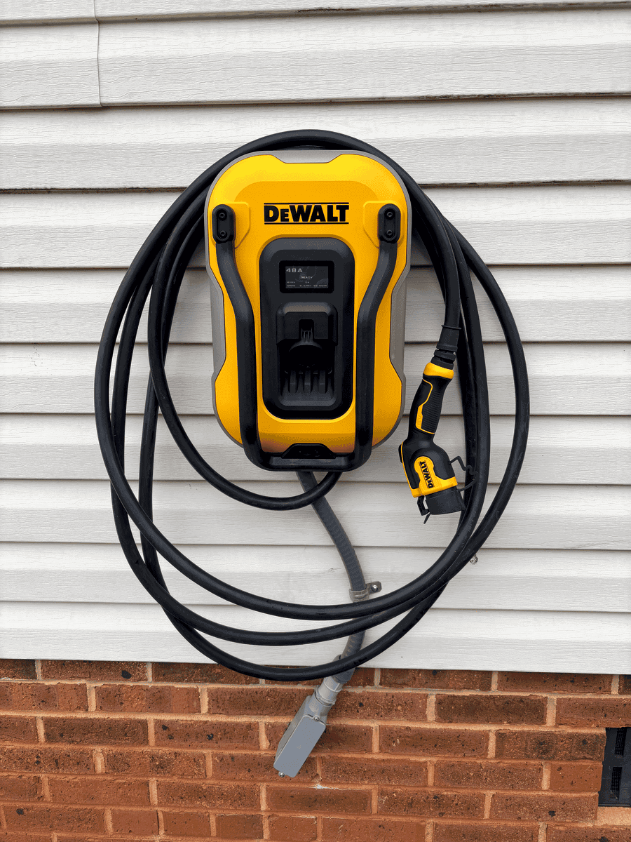 Yellow and black DeWalt outdoor electric vehicle charging station mounted on a white wooden wall, with a coiled charging cable attached. Demonstrating electric vehicle home charger installation services.