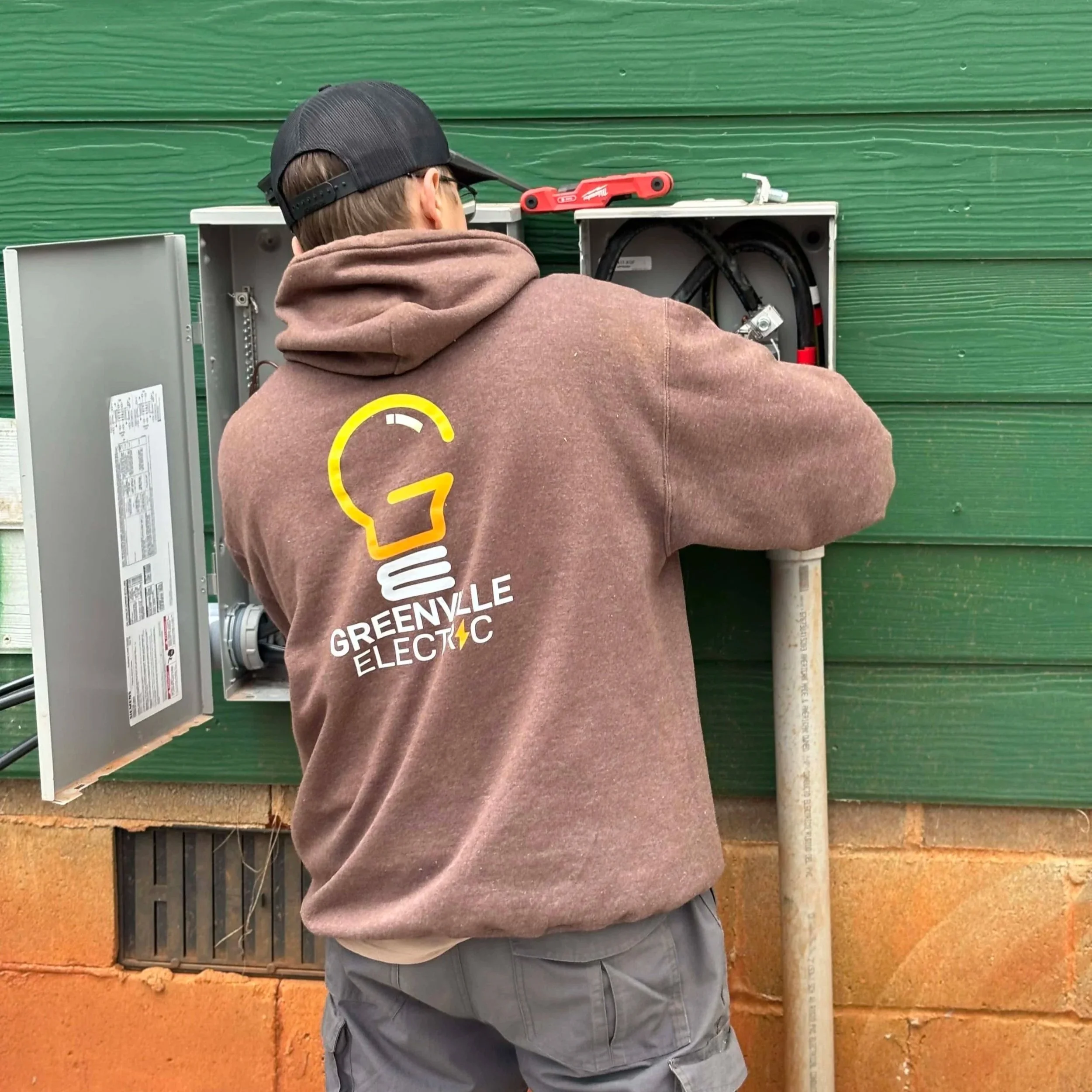 A person working on an outdoor electrical box mounted on a green wall, wearing a brown Greenville Electric hoodie and a black cap. Demonstrating generator transfer switch installation  and generator hookup services.