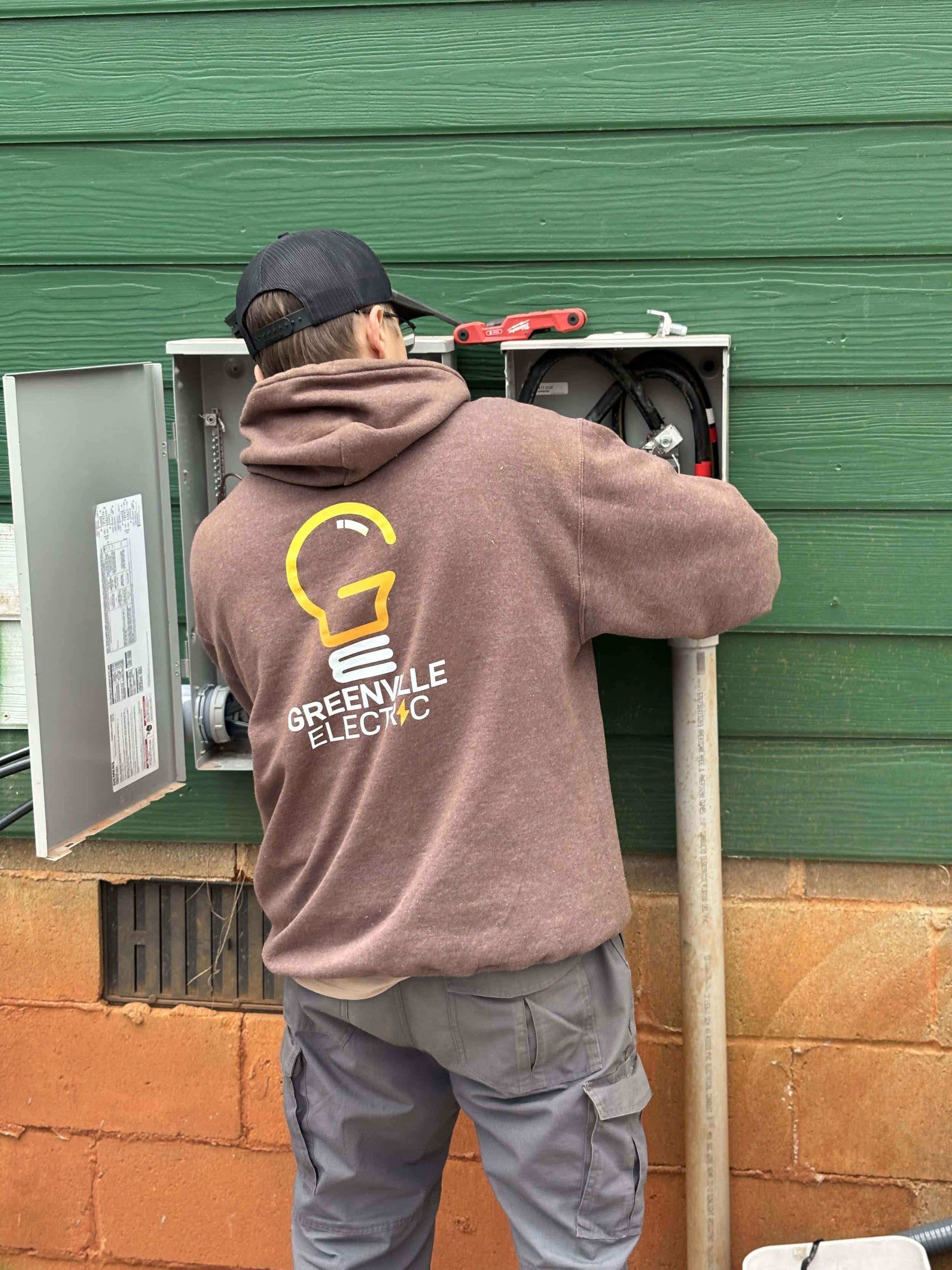 A worker in a brown hoodie with Greenville Electric logo installs or repairs an outdoor electrical panel on a green building wall.