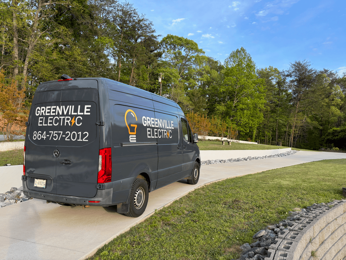 A dark gray delivery van with Greenville Electric branding and contact information, parked on a curved concrete sidewalk in a park with green grass, trees, and a clear blue sky.