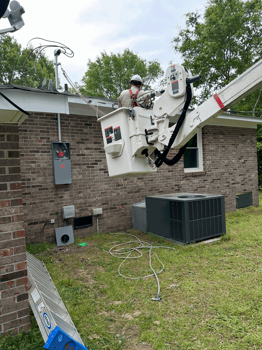 A repair technician in a bucket lift working on an electrical line outside a brick house, with tools and equipment on the ground. Demonstrating professional licensed electrician wiring.