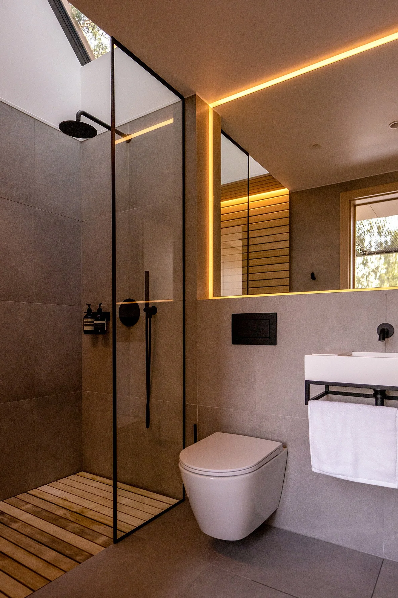Modern and efficient bathroom with walk-in shower with flush timber planks, wall-mounted toilet, sink, towel rack, and large skylight with trees outside, illuminated by warm recessed lighting.