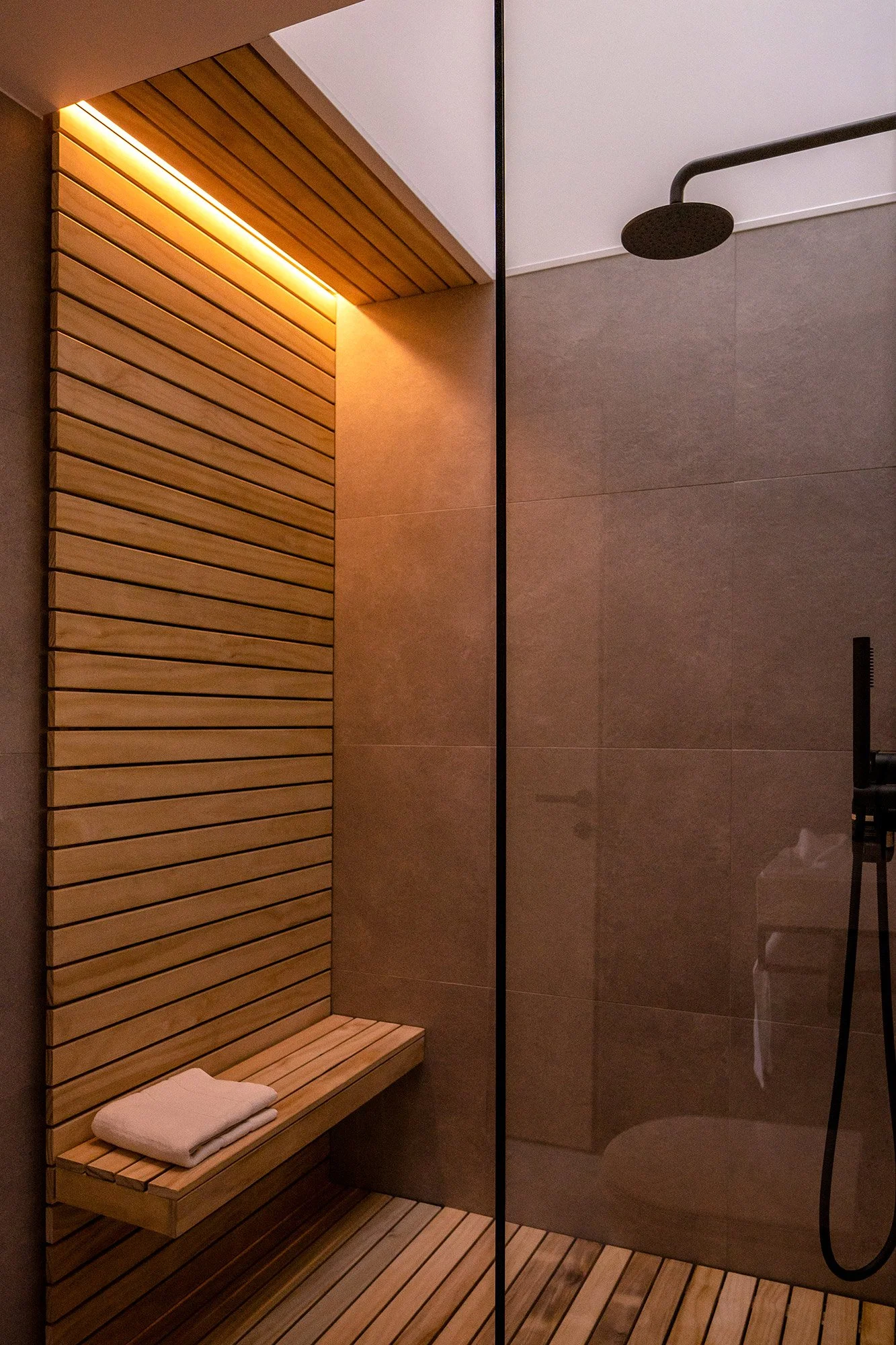 Interior of a modern shower with wooden slats, an integrated wooden bench for storing towels, tiled walls, a skylight and a rain shower head.