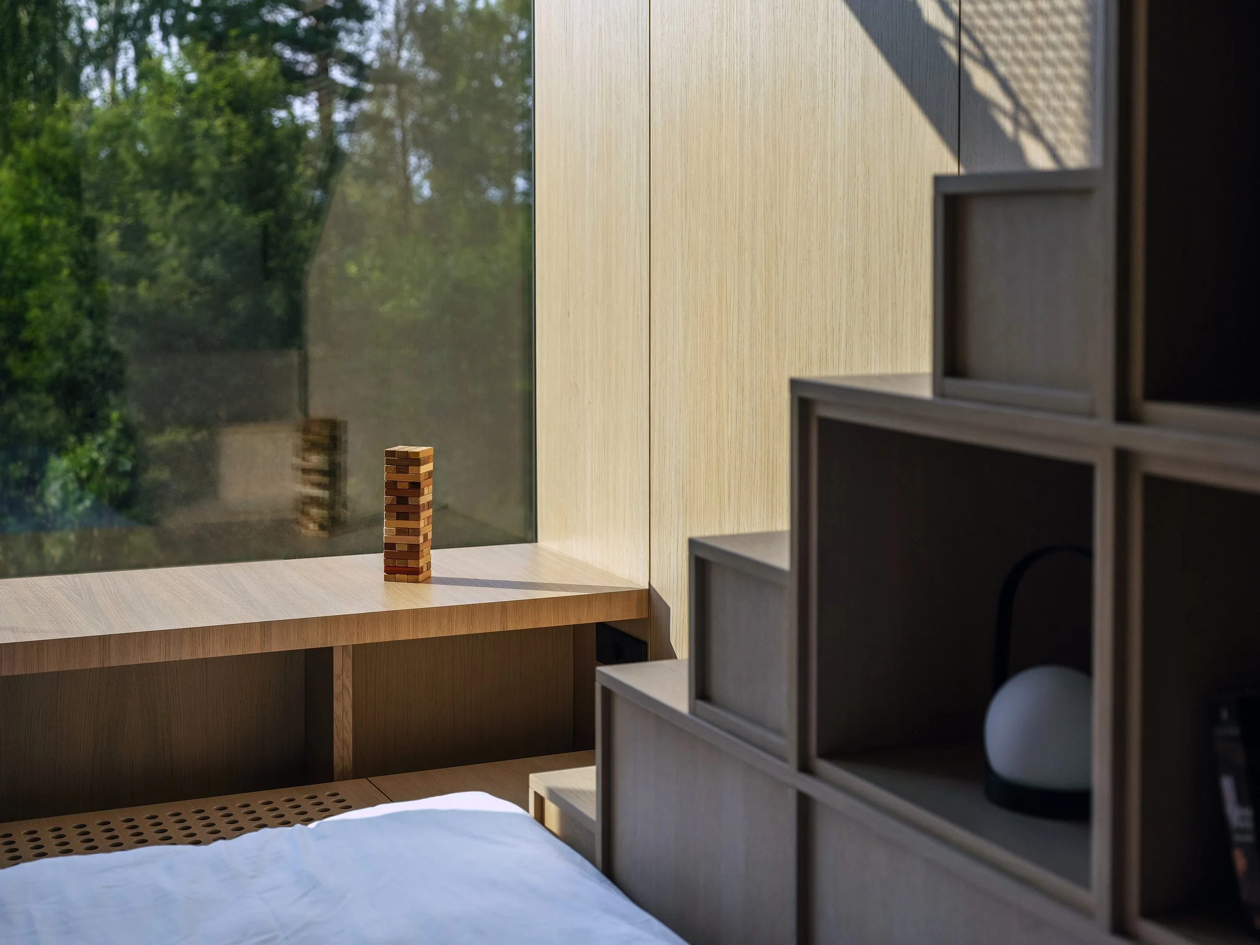 Modern bedroom with wooden furniture and a large window showing trees outside. A Jenga tower is on the window sill, and part of a bed with white sheets is visible in the foreground.