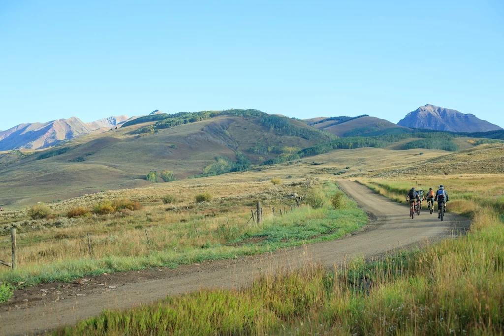 Three cyclists riding on a dirt road through a scenic grassy countryside with mountains visible in the background.