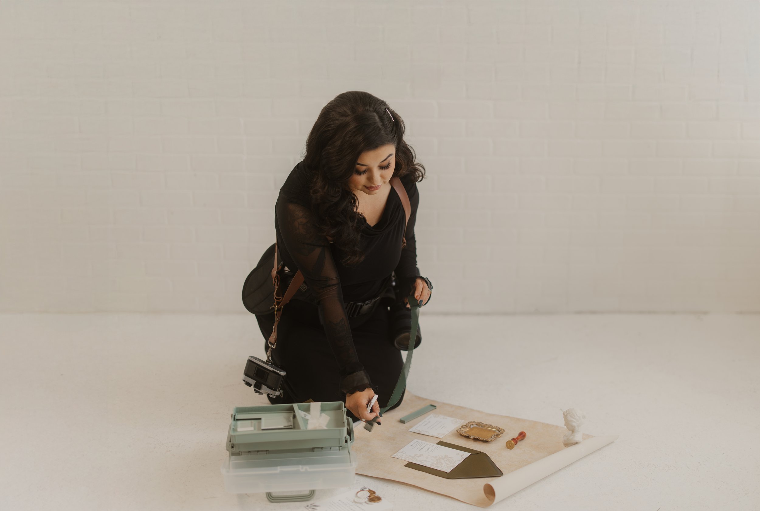 A woman dressed in black is kneeling on the floor surrounded by documents, a magnifying glass, small sculptures, and a camera. She appears to be examining or working on a project related to art or design.