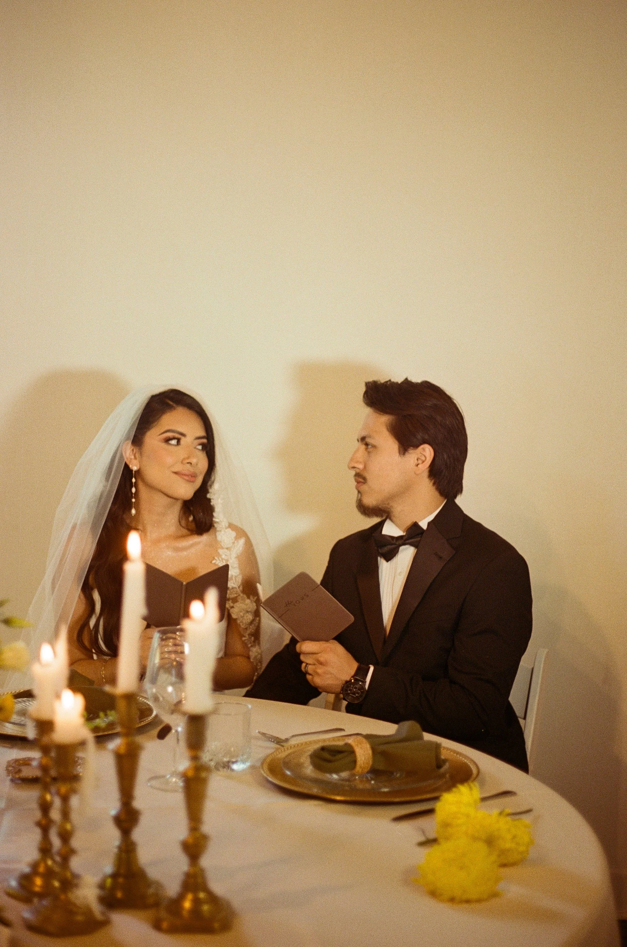 A bride and groom at their wedding ceremony, sitting at a table with candles and tableware, holding small booklets.
