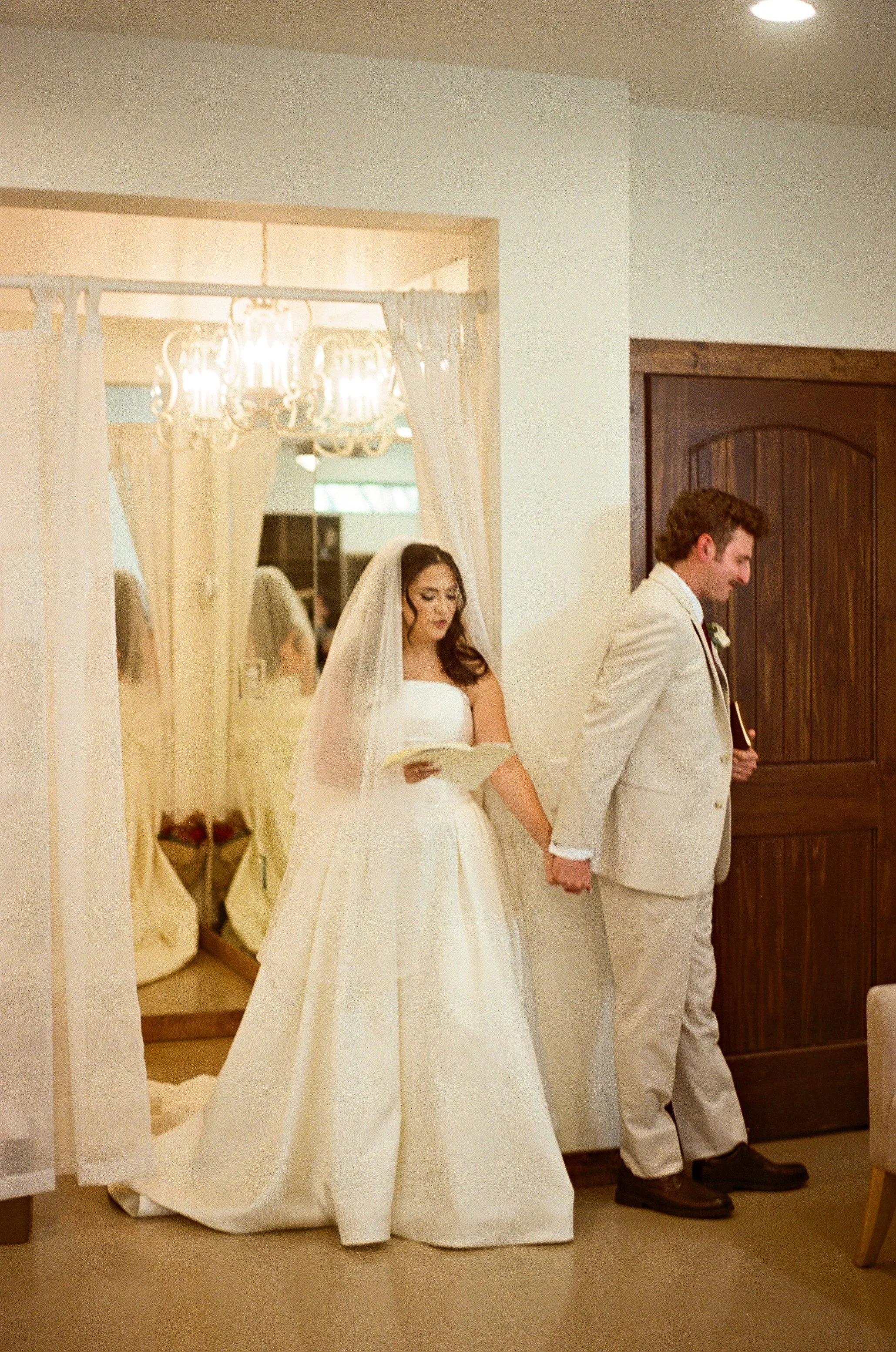 A bride holding hands with a groom during a wedding ceremony, standing in a room with white walls and a dark wooden door.
