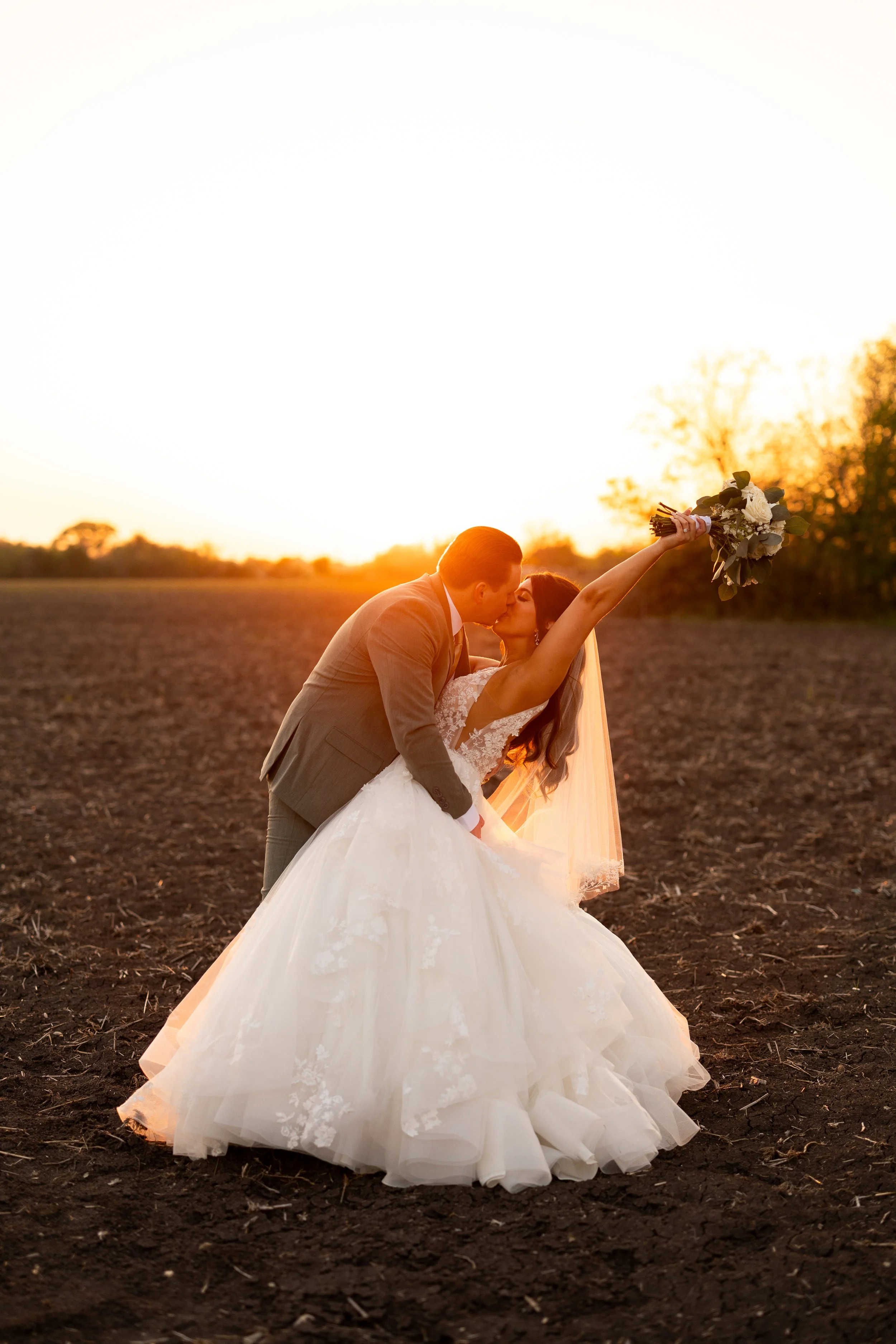 A newlywed couple sharing a kiss and holding a bouquet during sunset in a field.