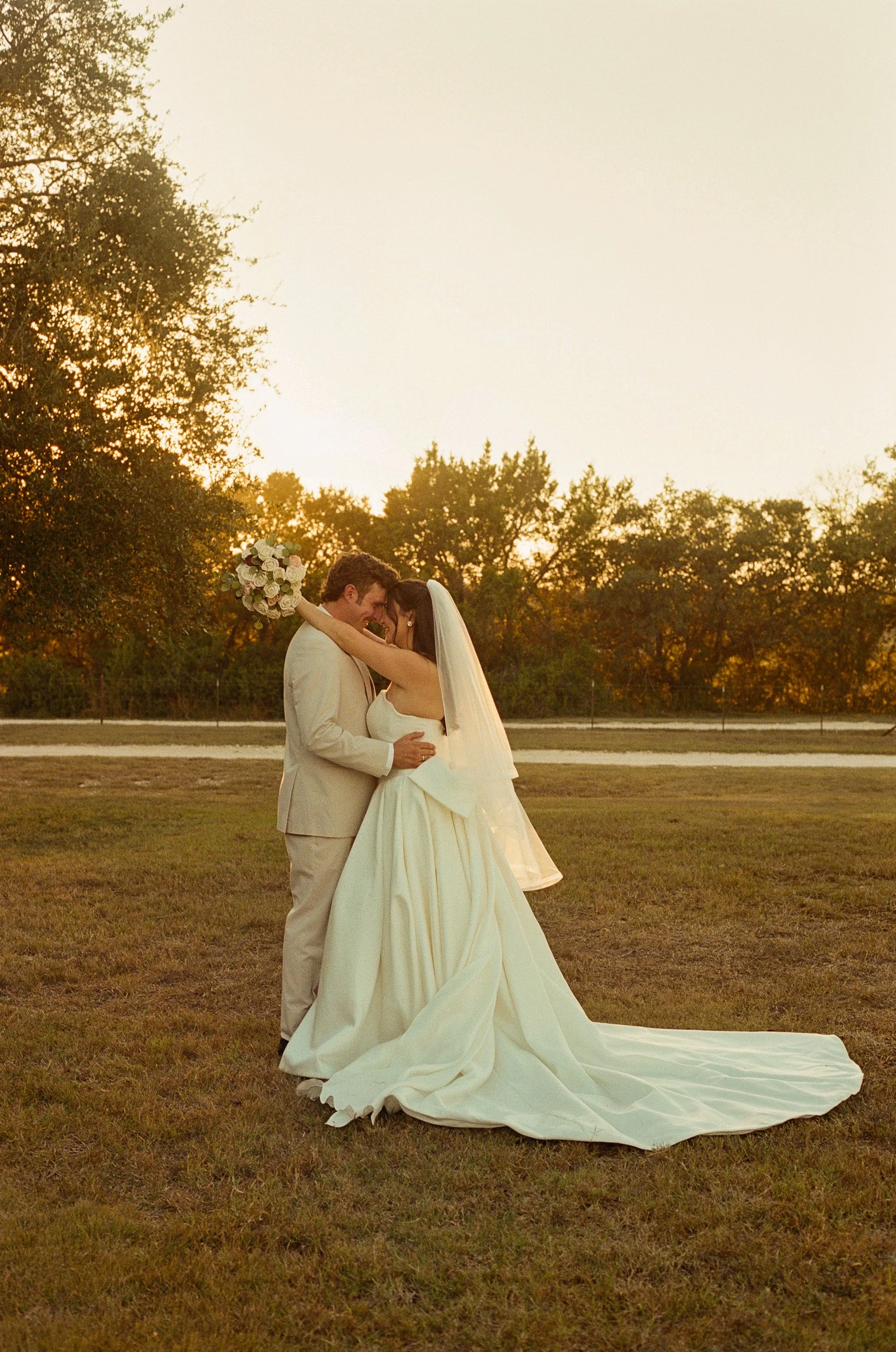 A bride and groom embrace in a field at sunset, with trees in the background, celebrating their wedding.