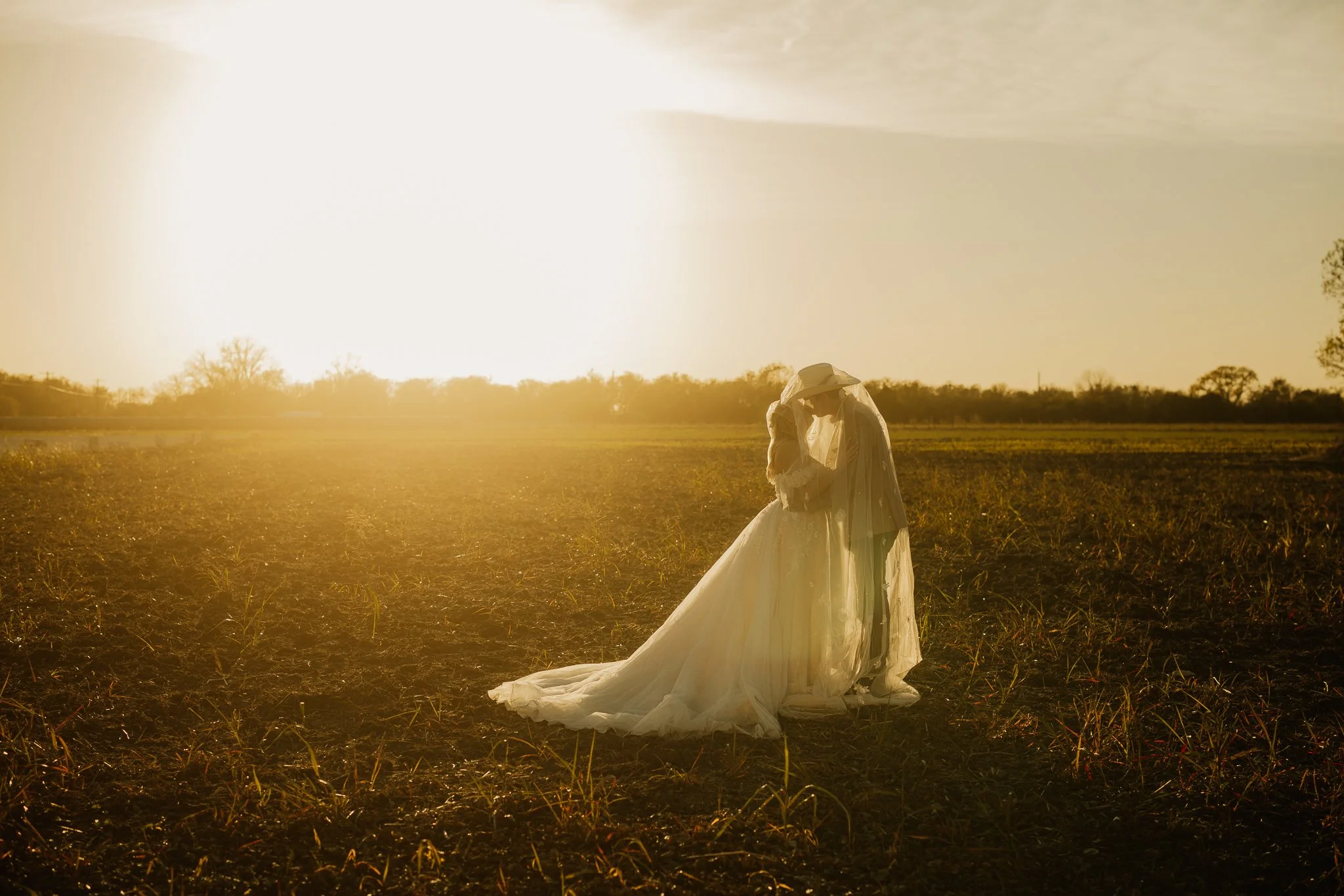 A bride in a wedding gown and veil standing in an open field with the setting sun in the background.