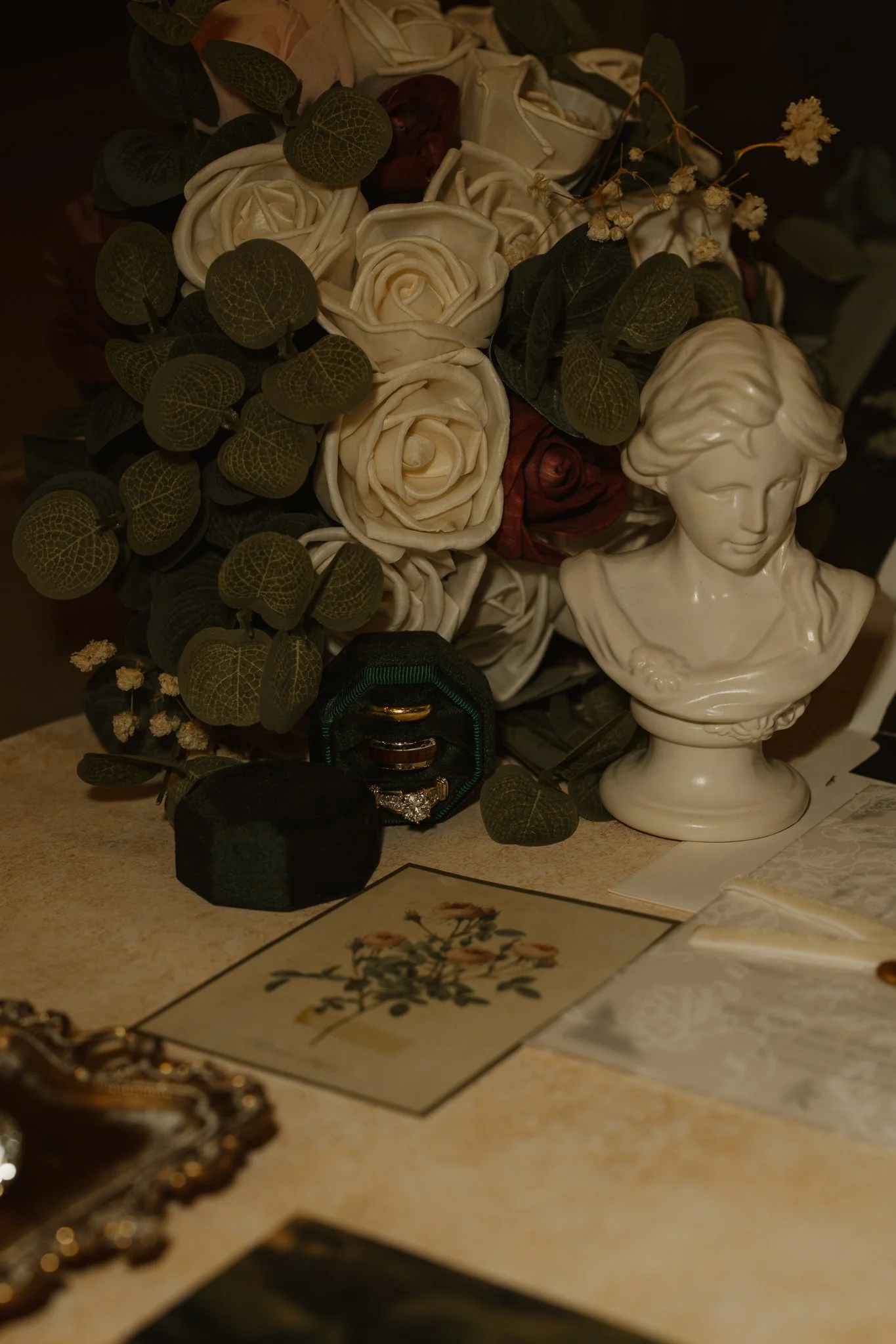 A still life arrangement with artificial white roses, green foliage, a white bust sculpture of a young girl, a jewelry box with rings, and vintage photos on a beige surface.