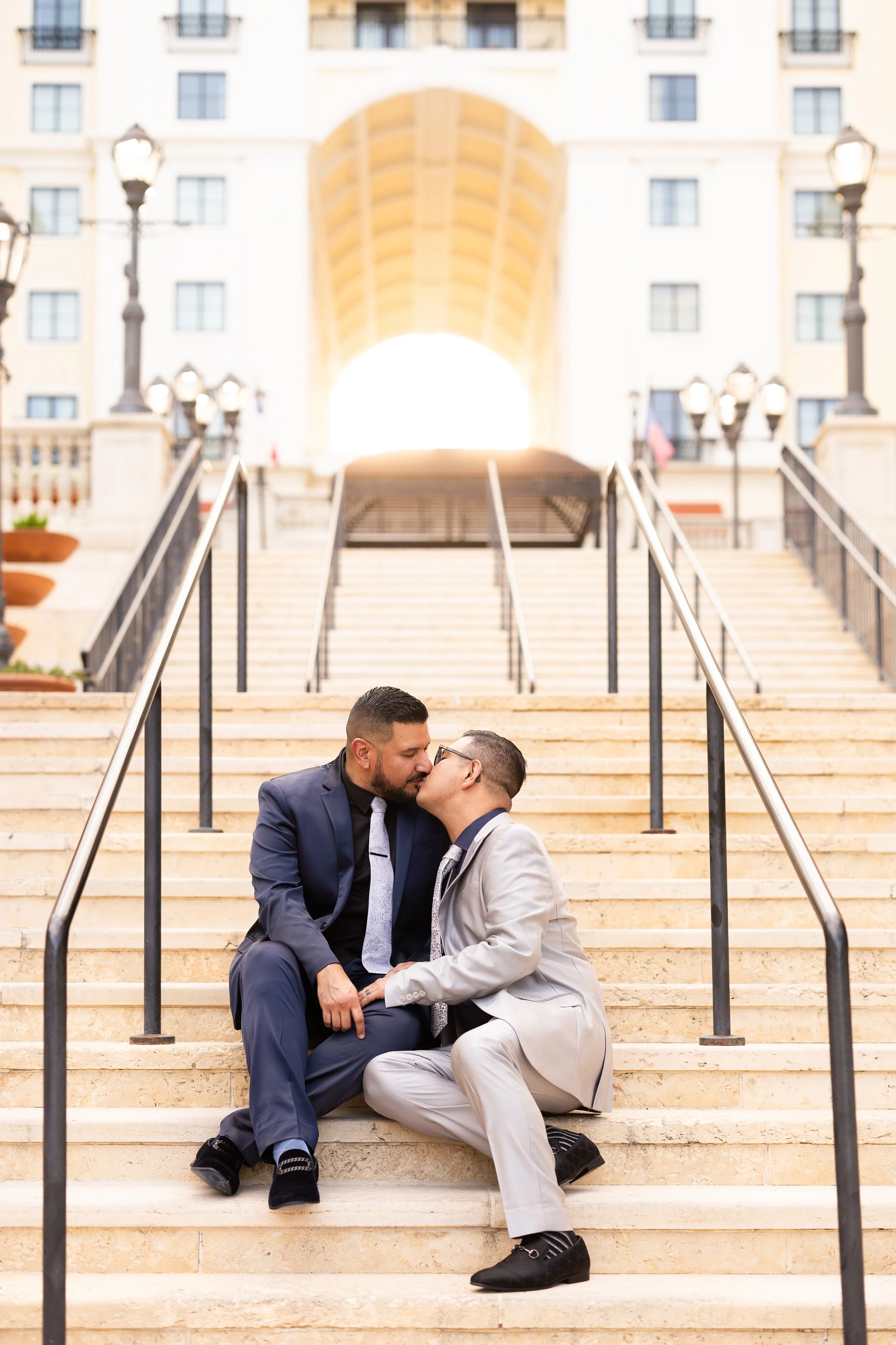 Two men in suits sharing a kiss while sitting on steps outside a building with an archway and cityscape