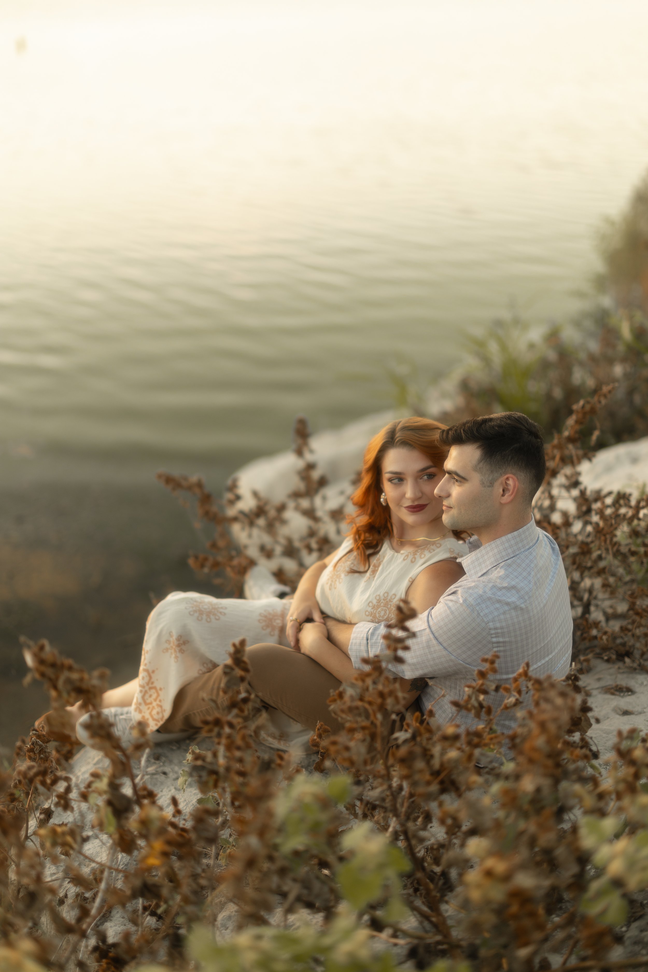 A young couple sitting on the ground by a body of water, surrounded by dry plants, during sunset.