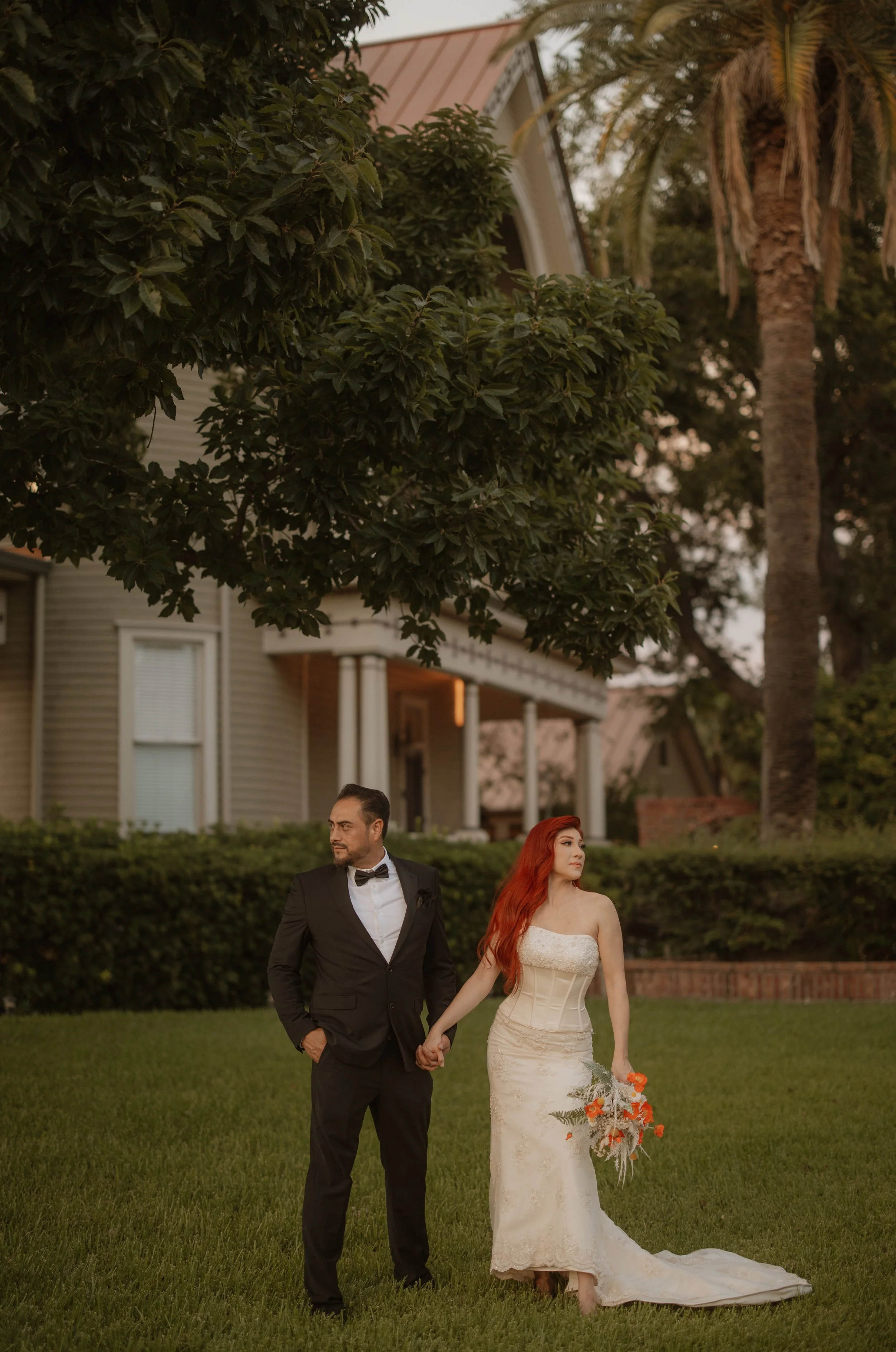 A couple dressed in wedding attire standing outdoors on a lawn, holding hands, with a large tree and a house in the background.