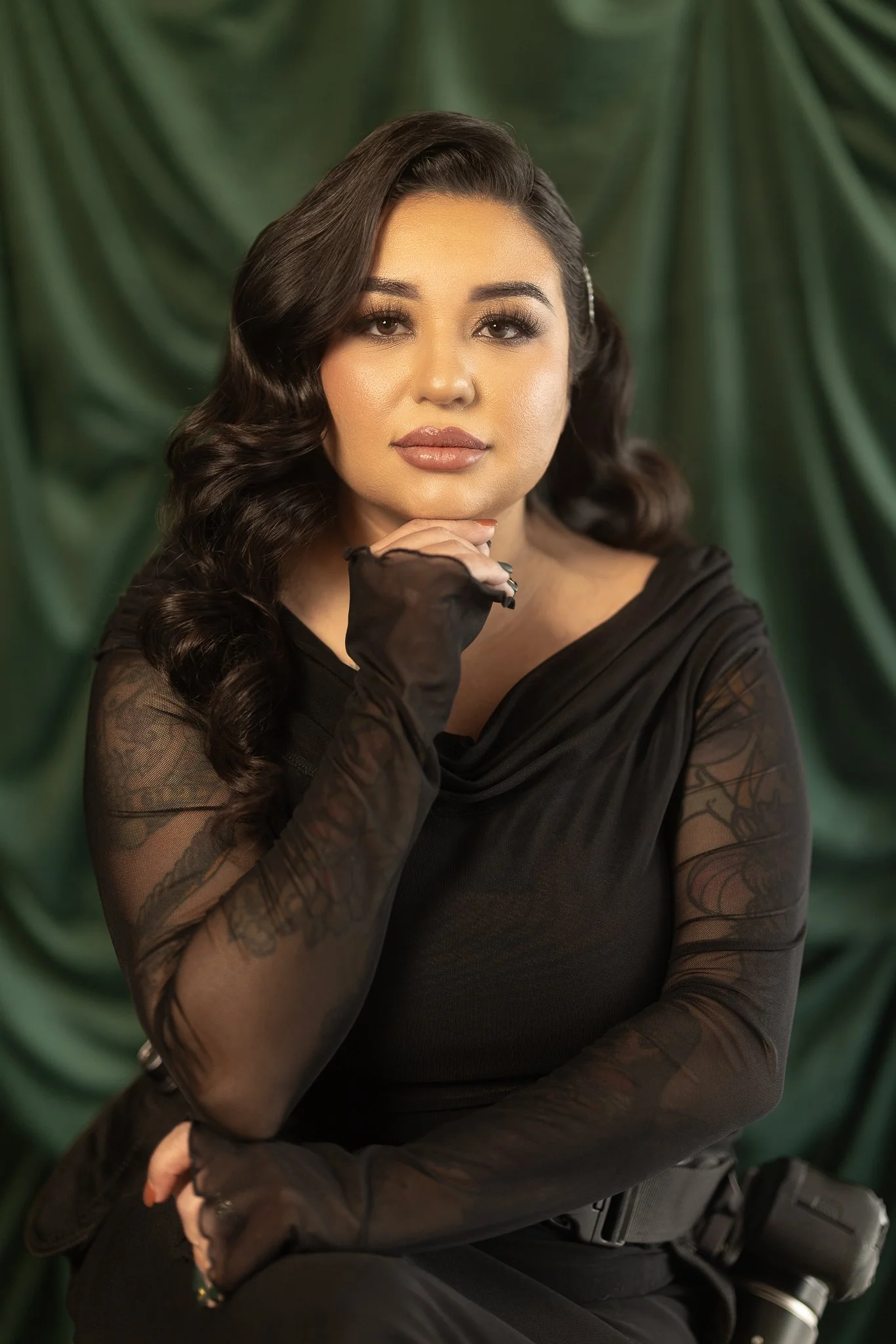 Woman with dark wavy hair wearing a black sheer long-sleeve top, posing with hand near face, against green draped background.
