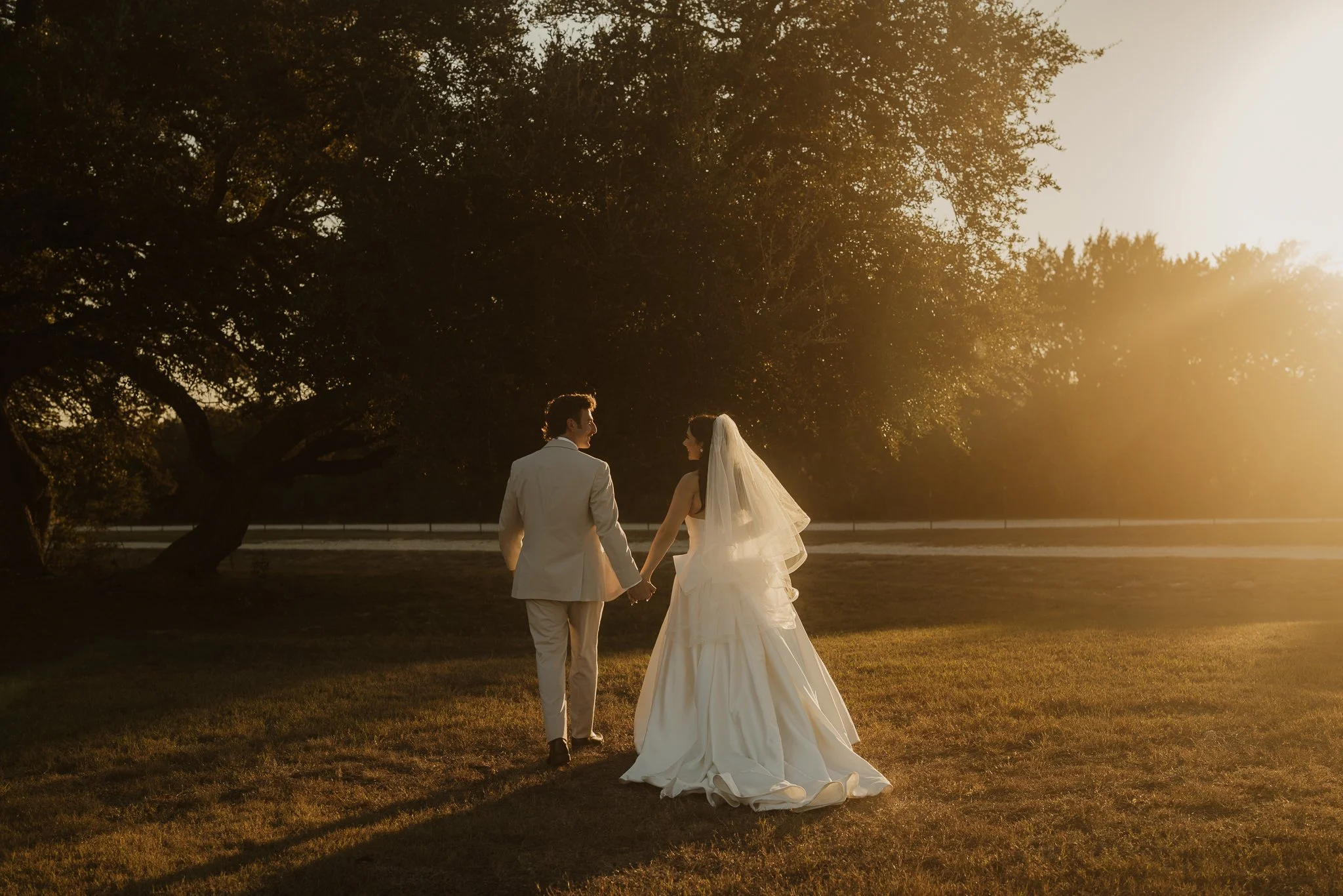 A bride and groom holding hands and walking in a grassy field during sunset, with trees in the background.