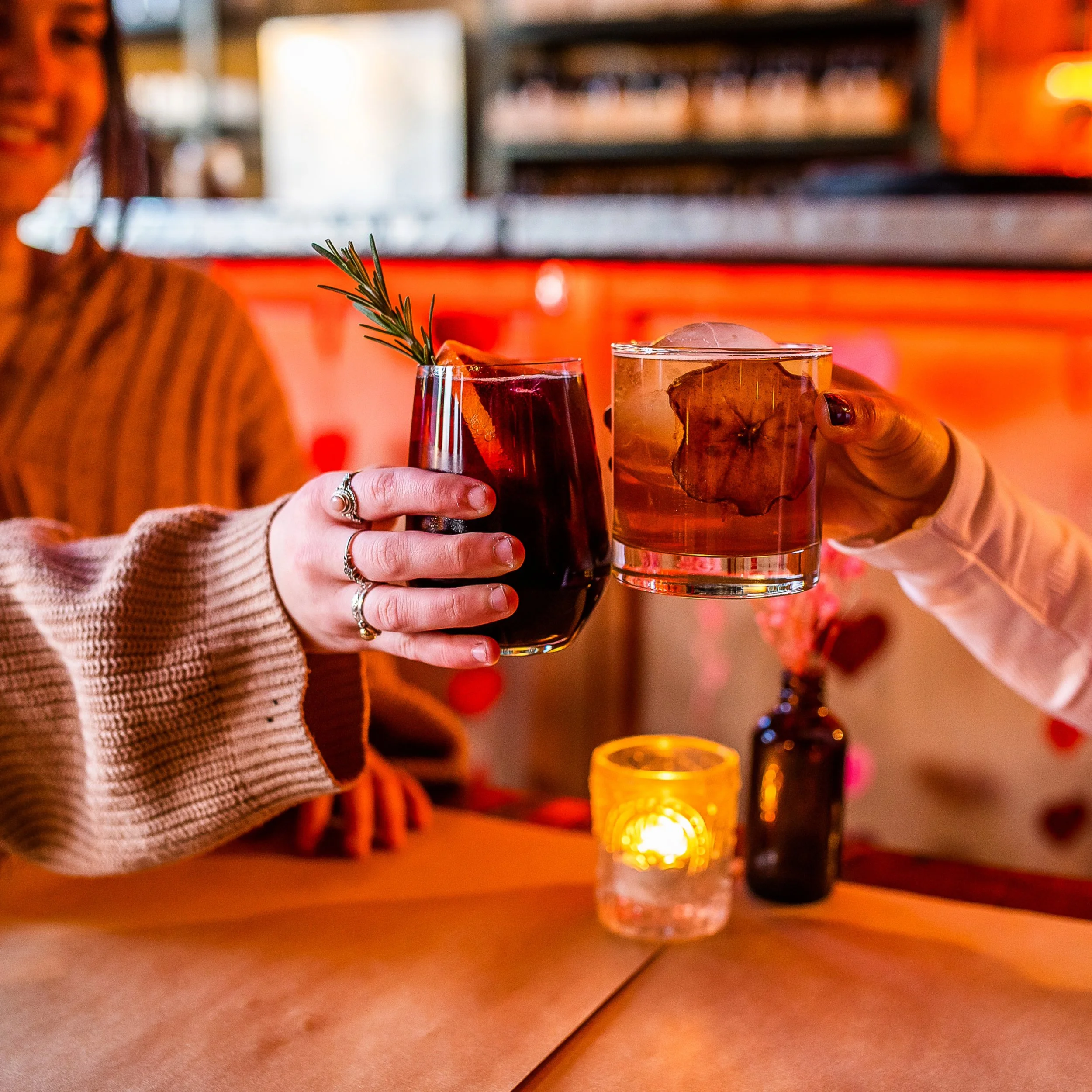Two people hold up glasses with cocktails, one with a sprig of rosemary, in the cozy, warmly-lit candle and cocktail bar. 