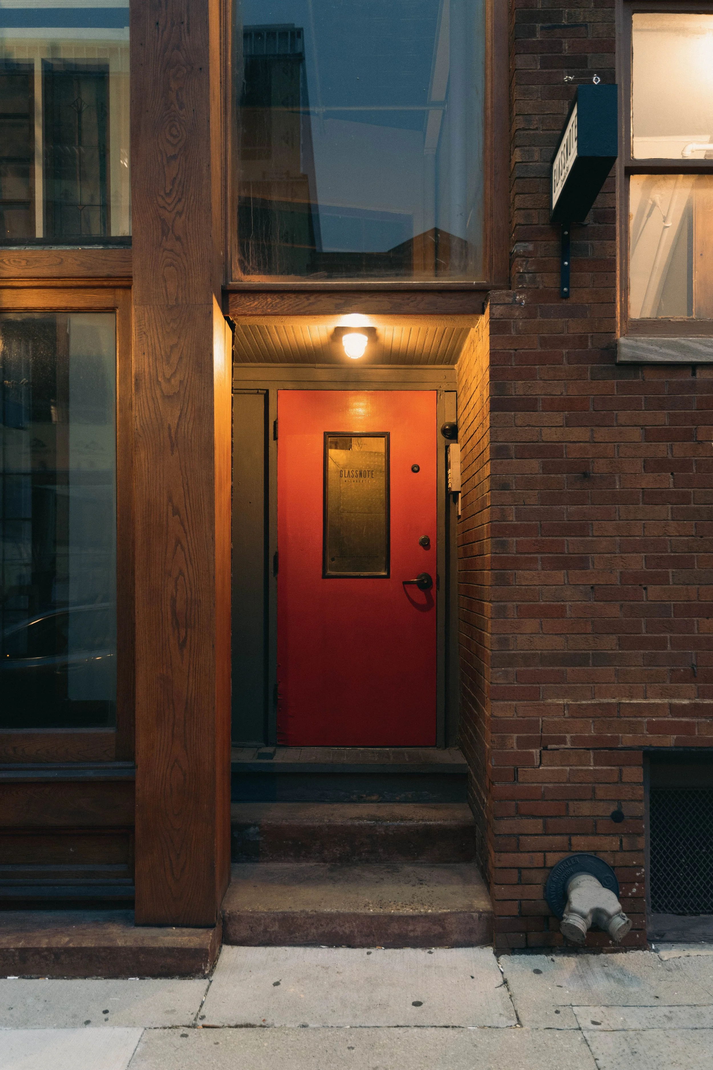 Close-up of the candle bar building entrance with a red door, a small window on the door labeled 'Glassnote,' brick and wood exterior, with a warm light overhead.
