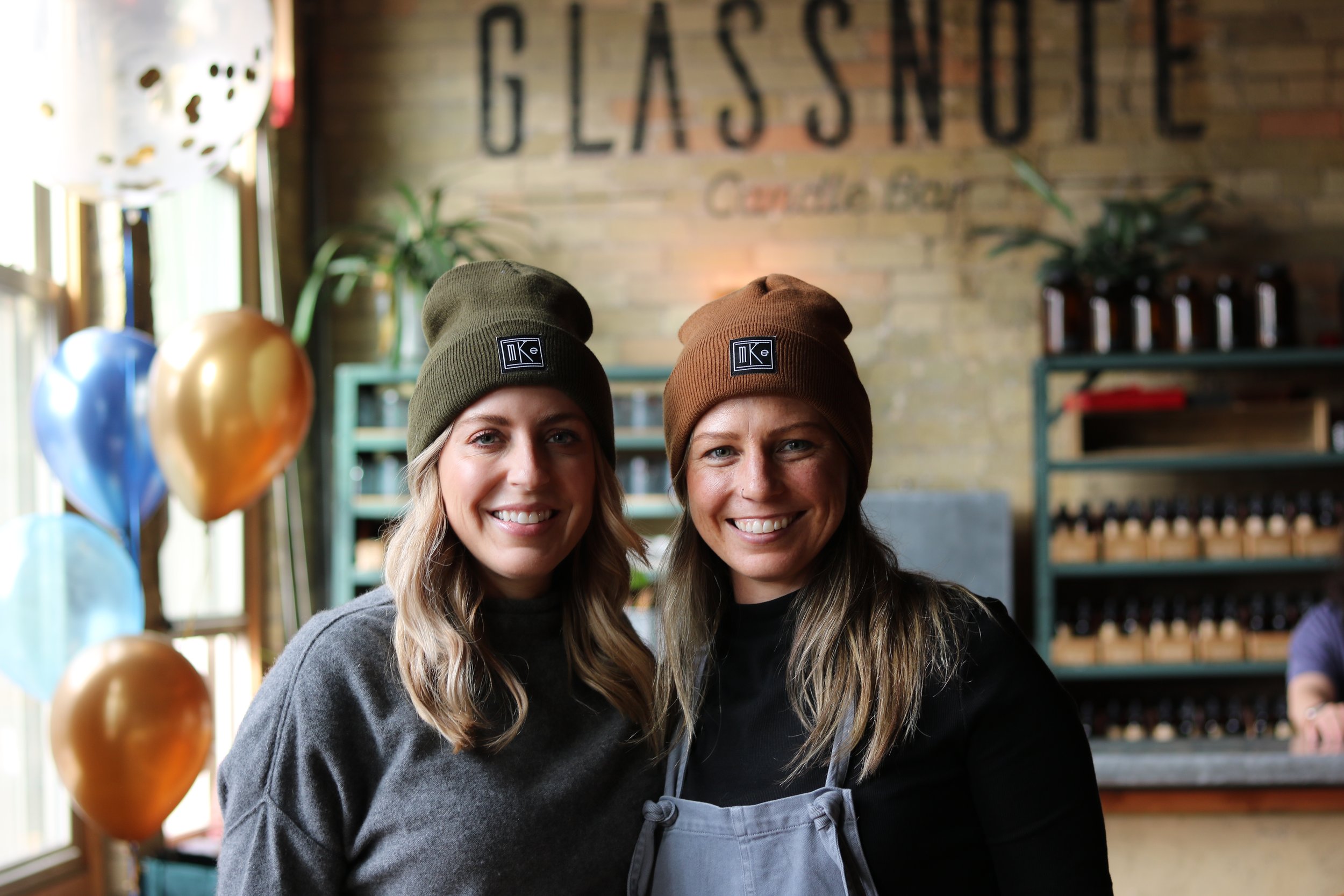 Two women smiling inside the candle bar, wearing beanies, standing in front of a brick wall with the word 'GLASSNOTE' on it, and decorative balloons in the background during a private event.