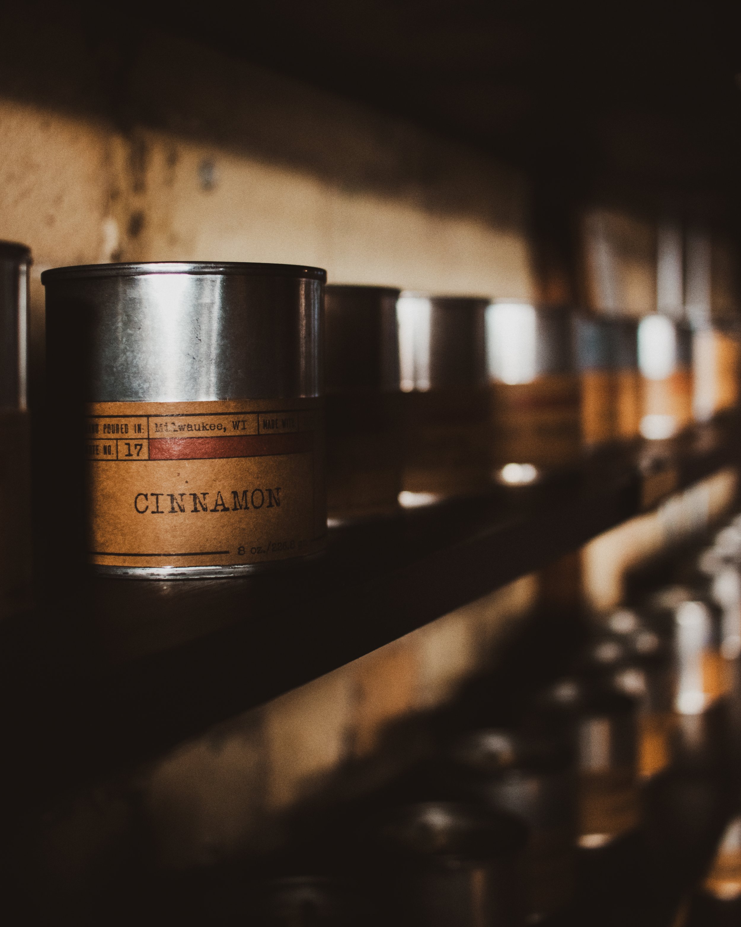 A row of metal fragrance cans on a black shelf, with one labeled 'CINNAMON' in the foreground, showcasing the fragrance wall.