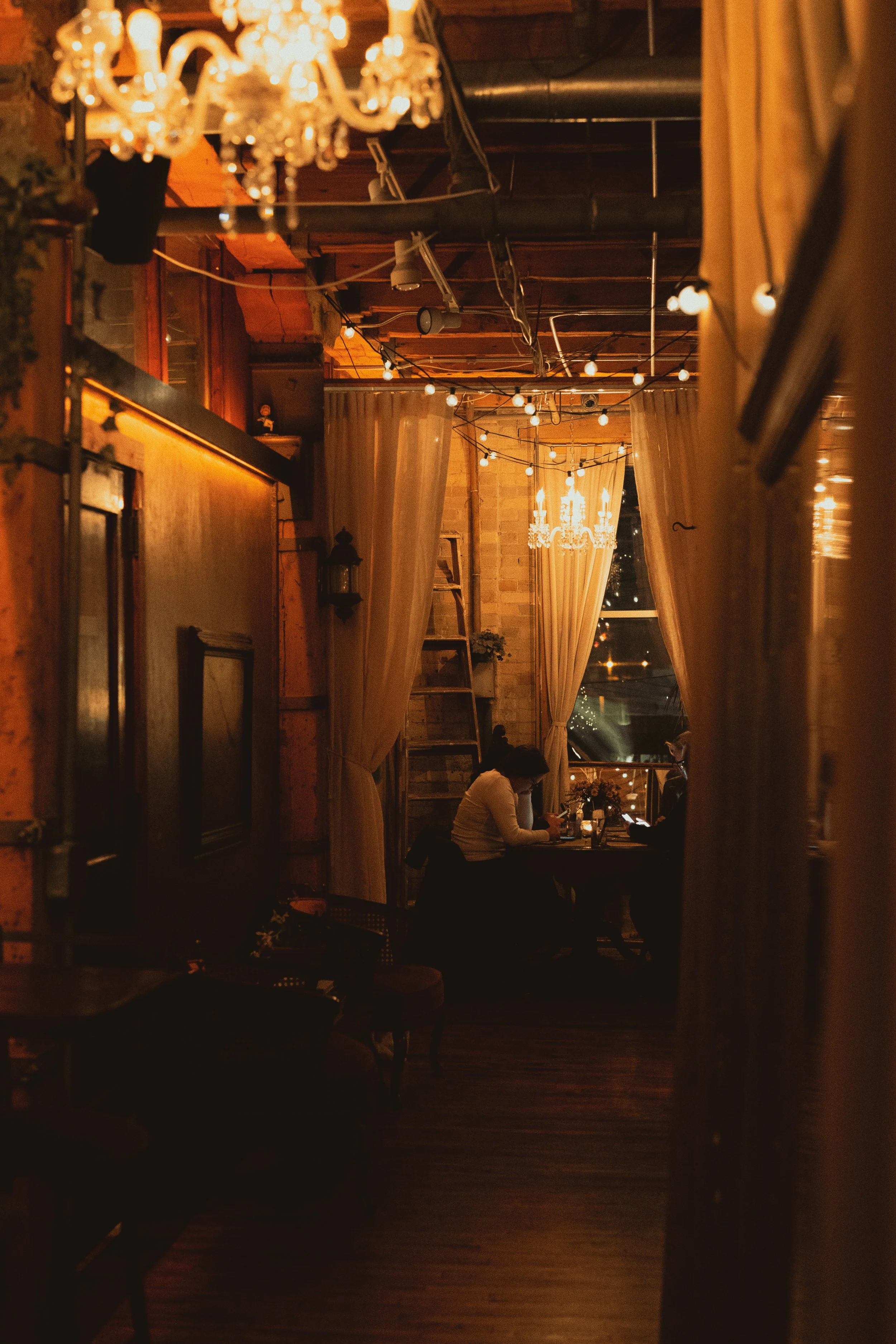 A cozy, dimly lit hall with warm ambient lighting, featuring a chandelier and string lights hanging from the ceiling. There are large curtains framing a window and cozy seating options.