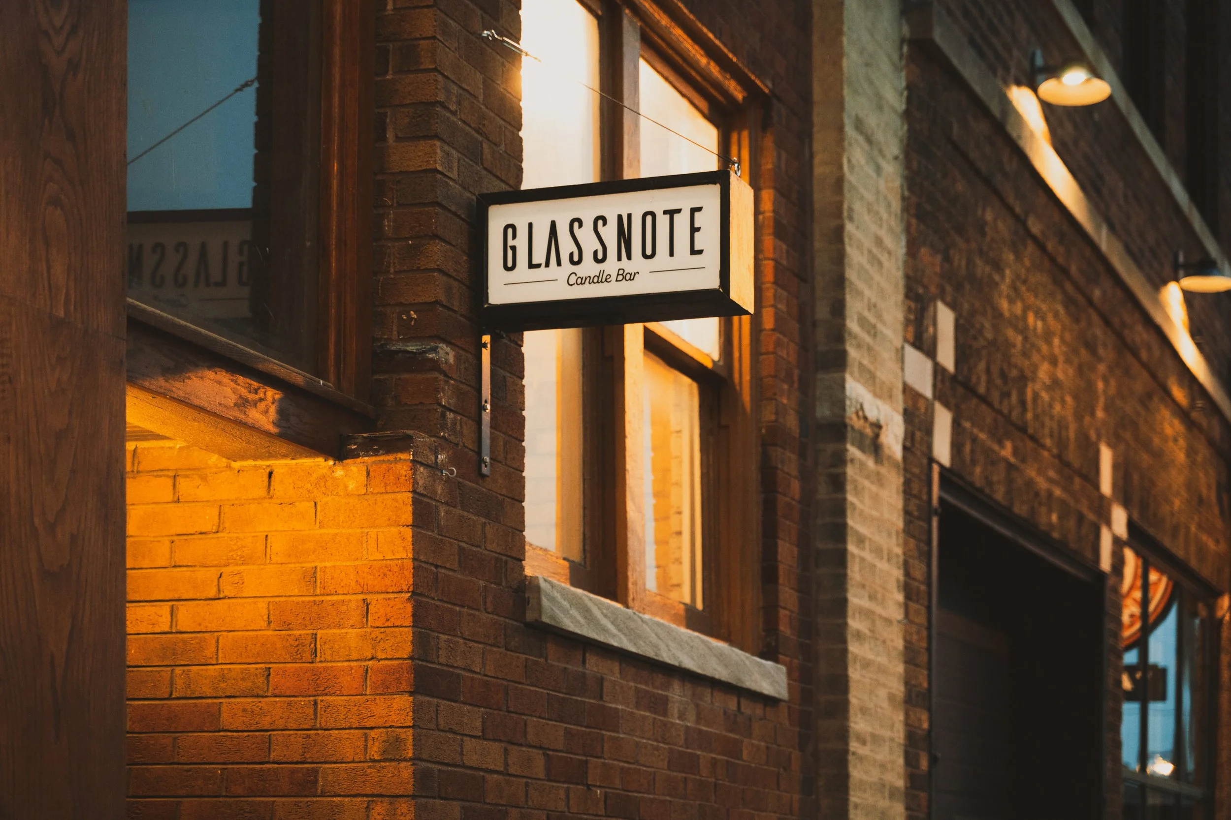 Sign for Glass Note Candle Bar hanging outside a brick building at sunset.