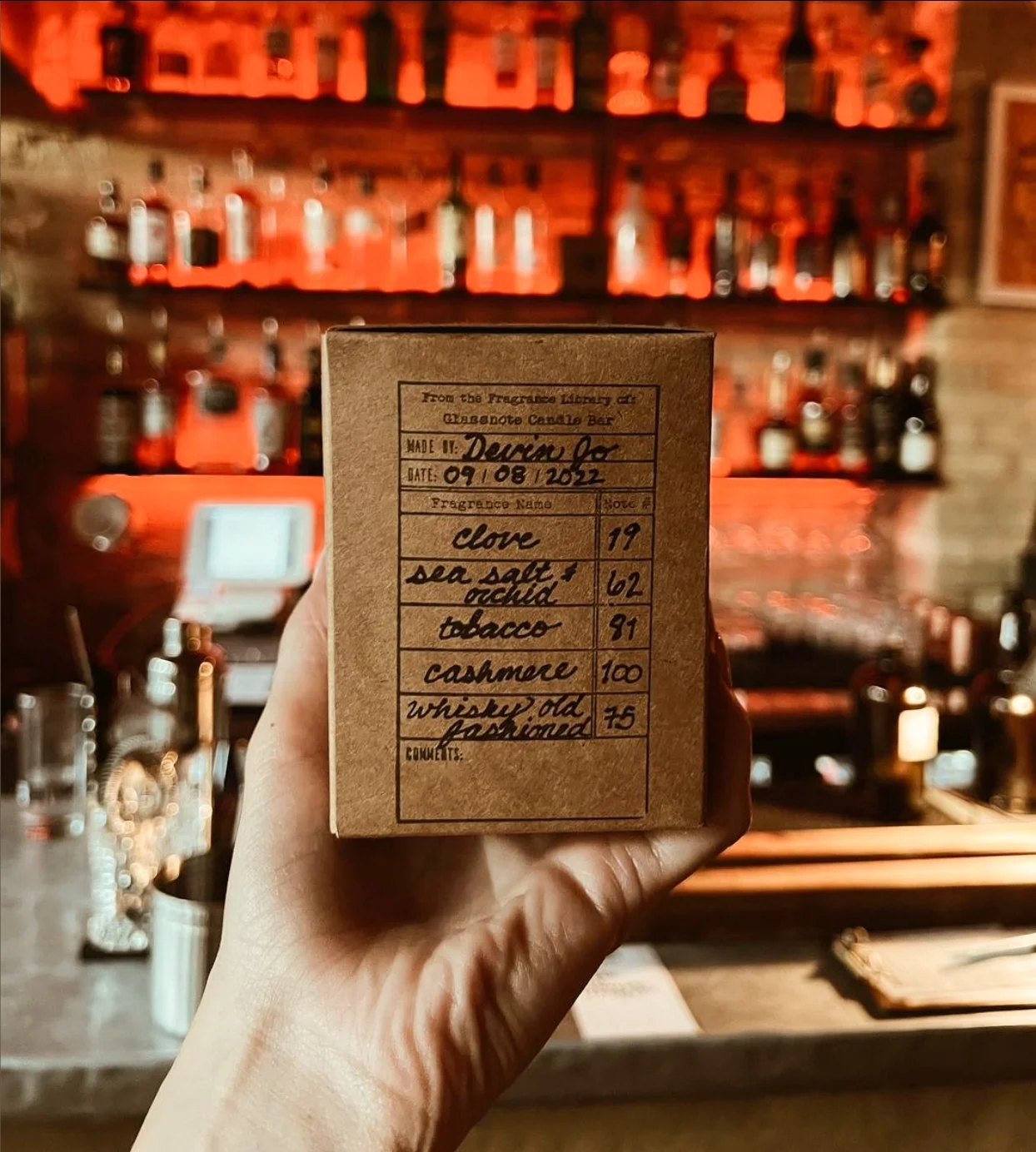 A person holding a small brown box with handwritten fragrance ingredients and details in glassnote candle bar. The background shows bottles on shelves and bar equipment with warm lighting.