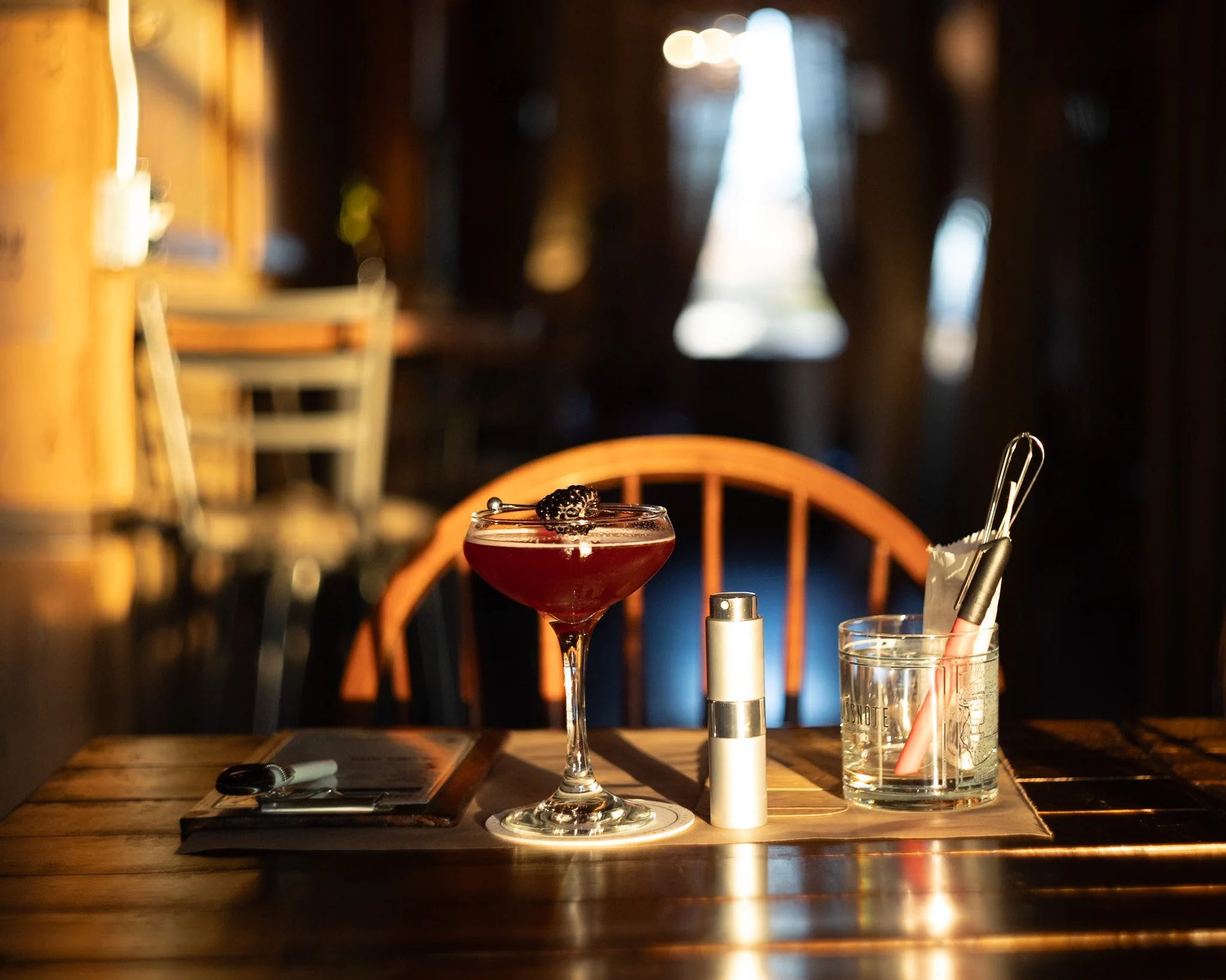 A wooden table with a cocktail glass filled with a red drink garnished with a blackberry, a small silver container, a glass holding a straw, black tongs, and a notepad with a pen against a dimly lit background.